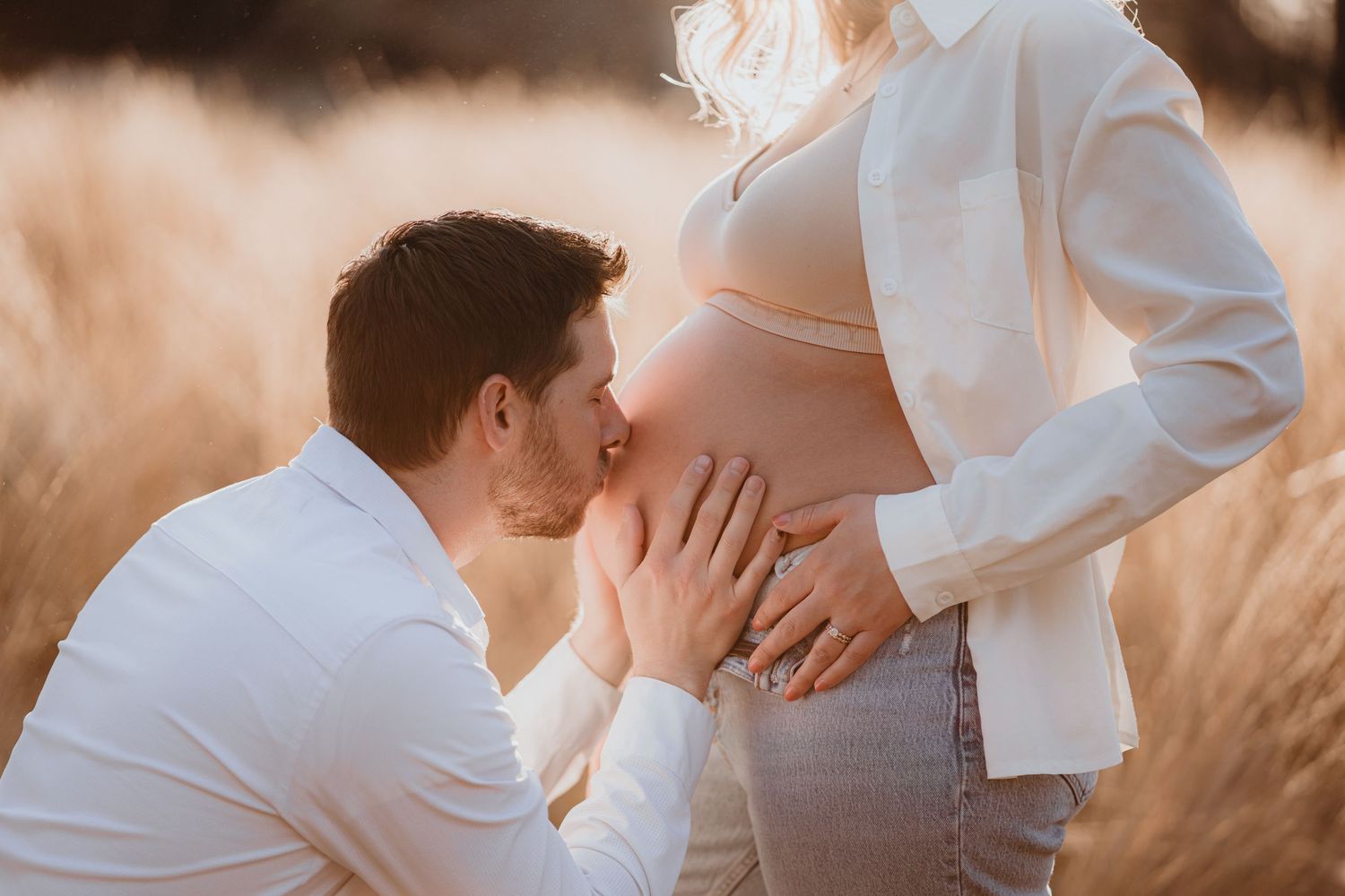 Man lovingly kissing pregnant woman's bare belly in golden field at sunset.
