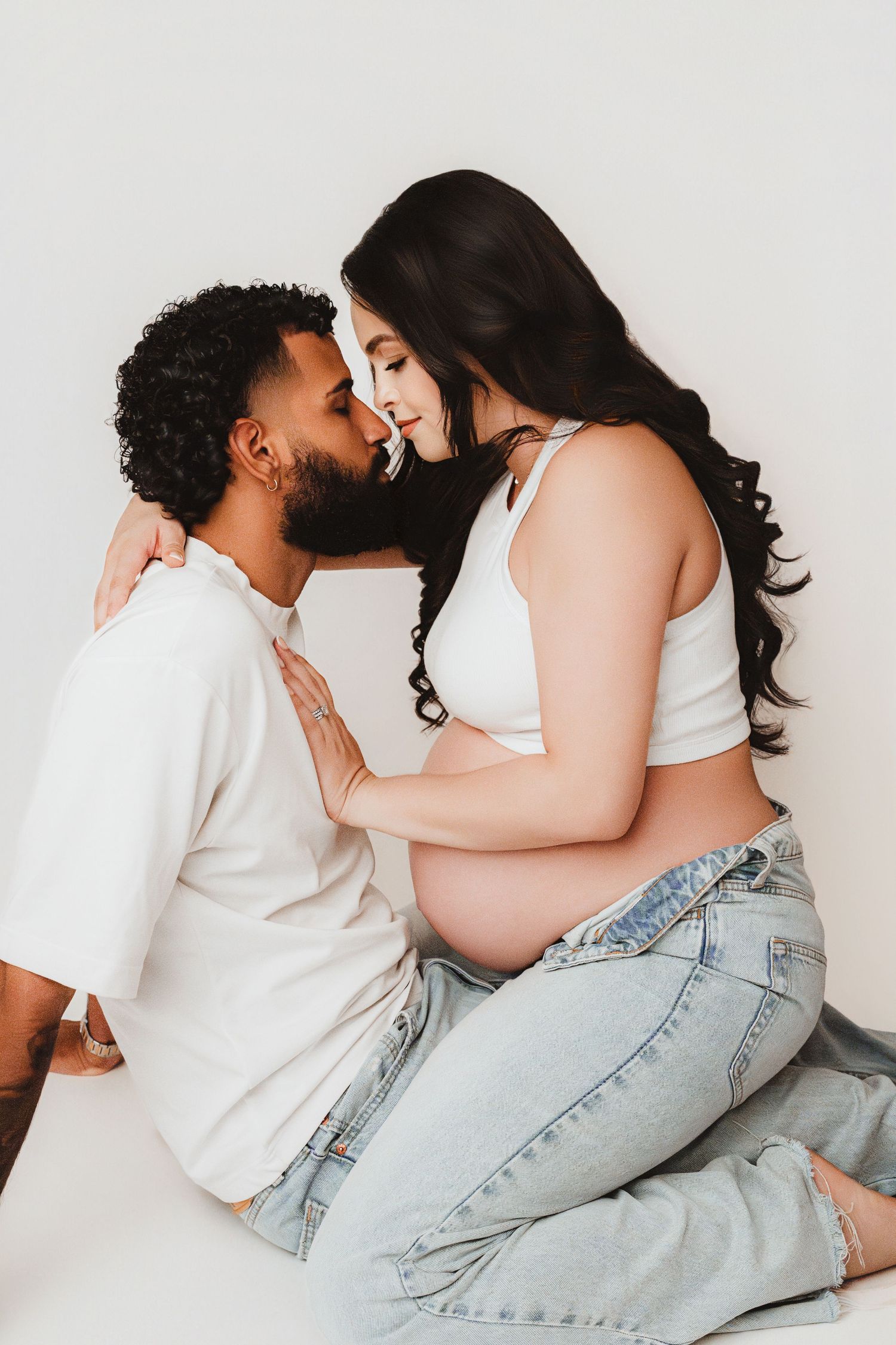Expecting couple almost kissing, woman cradling baby bump in white crop top and jeans.