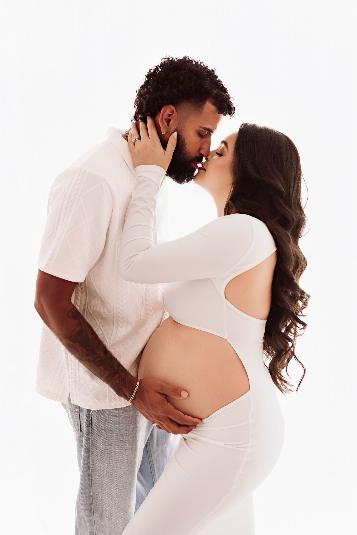 Intimate maternity portrait of interracial couple kissing, pregnant woman in white cutout outfit cradling her bare bump.