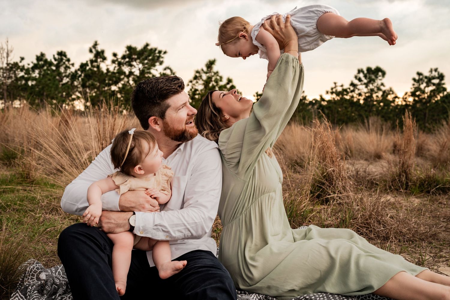 Mother lifting baby in the air while father holds toddler on a blanket in a golden outdoor field.