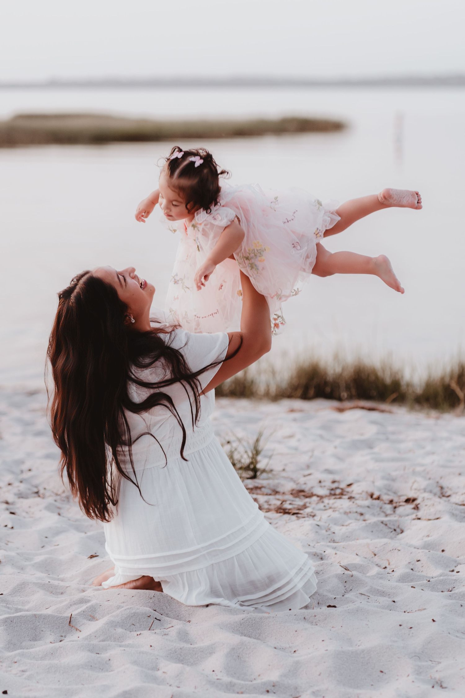 Joyful mother lifting laughing baby girl in pink dress into the air on beach.