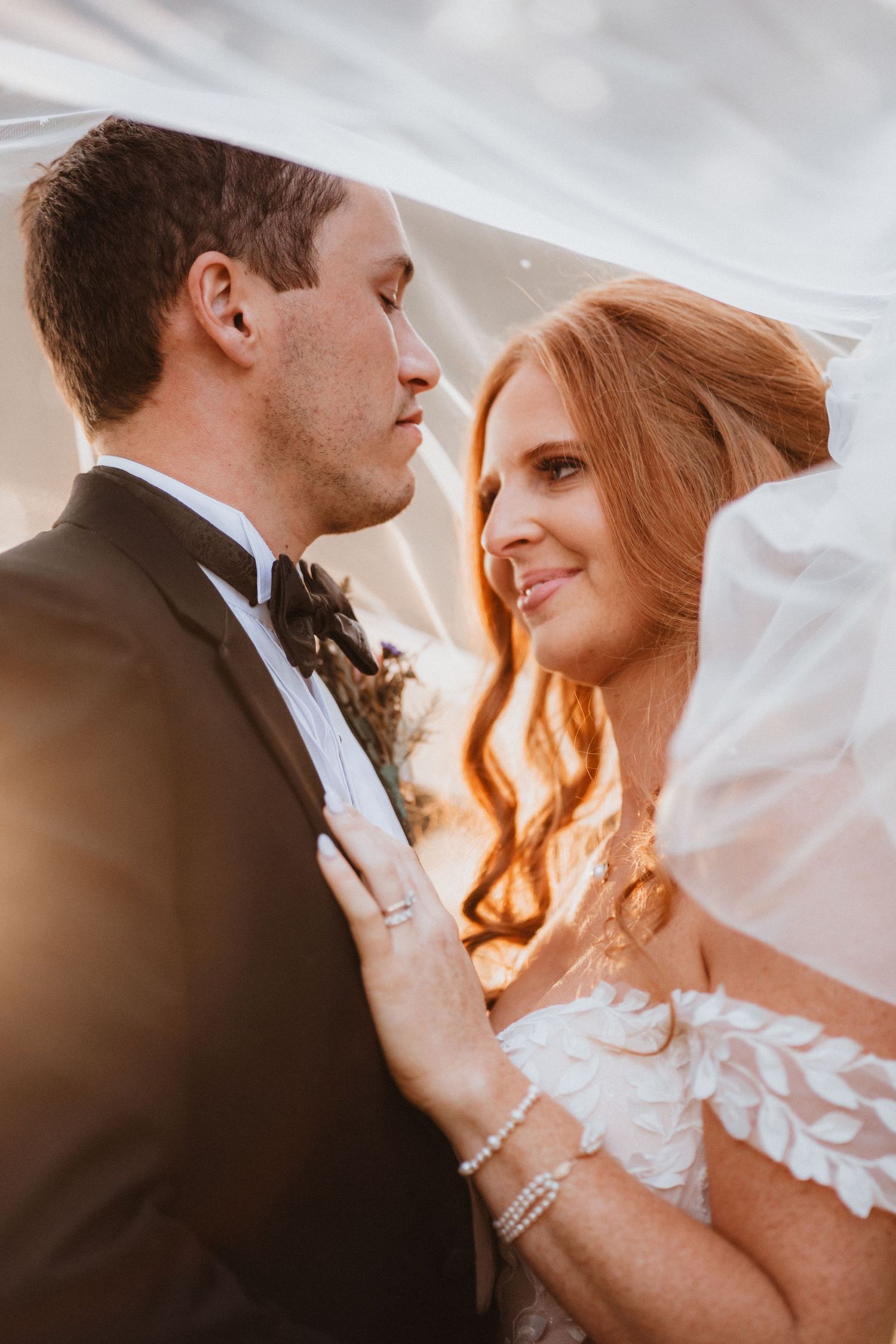 Bride and groom share an intimate moment under a veil in golden sunlight, bride smiling in lace gown.
