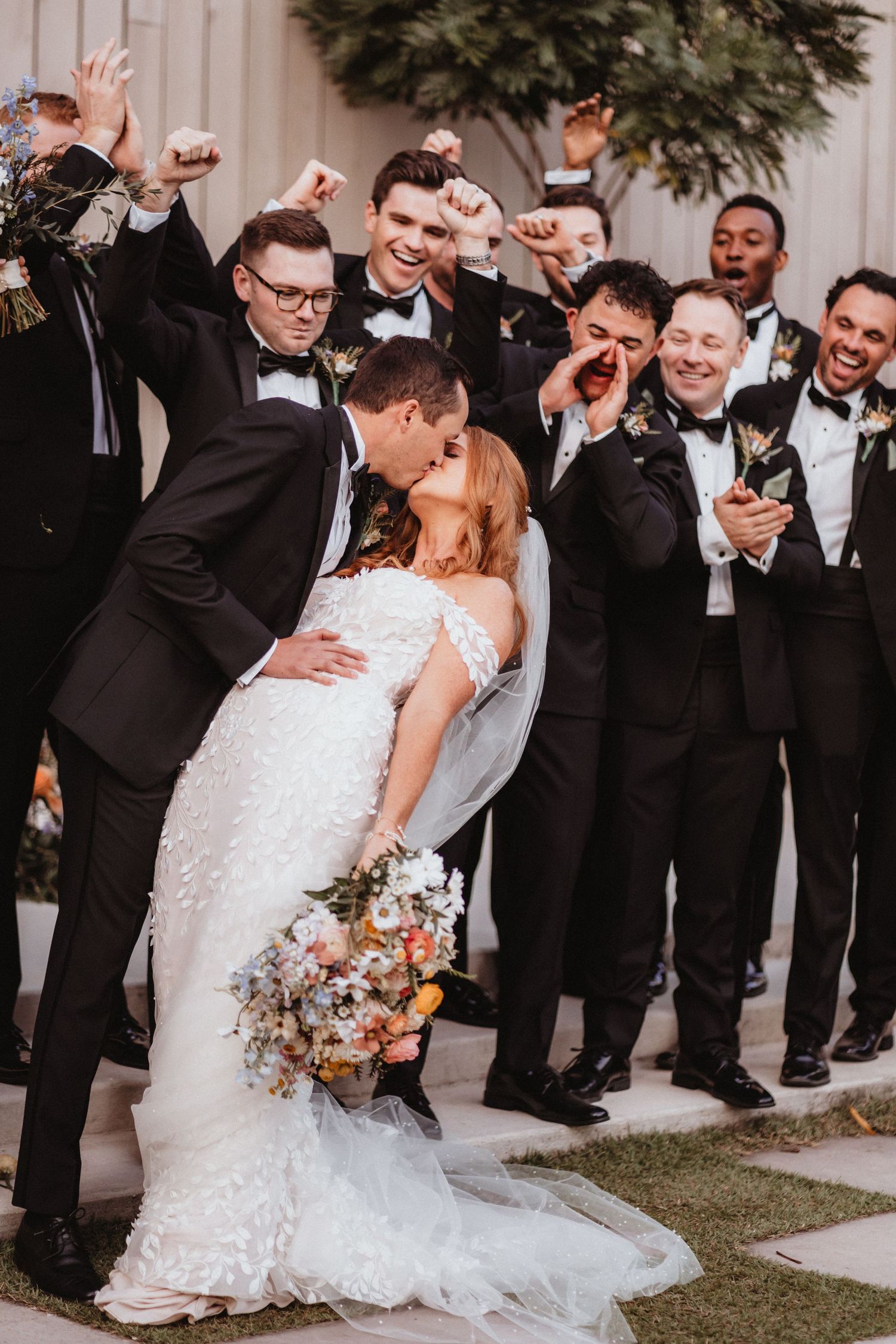 Groom dips bride for a kiss while groomsmen in black tuxedos celebrate enthusiastically behind them.