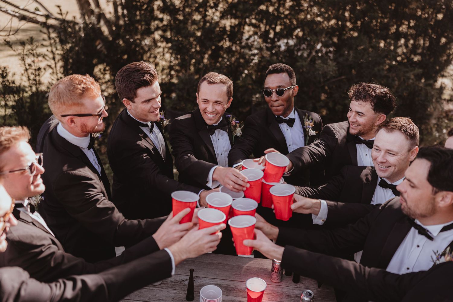 Groomsmen in black tuxedos toasting with red solo cups around a wooden table at a wedding.