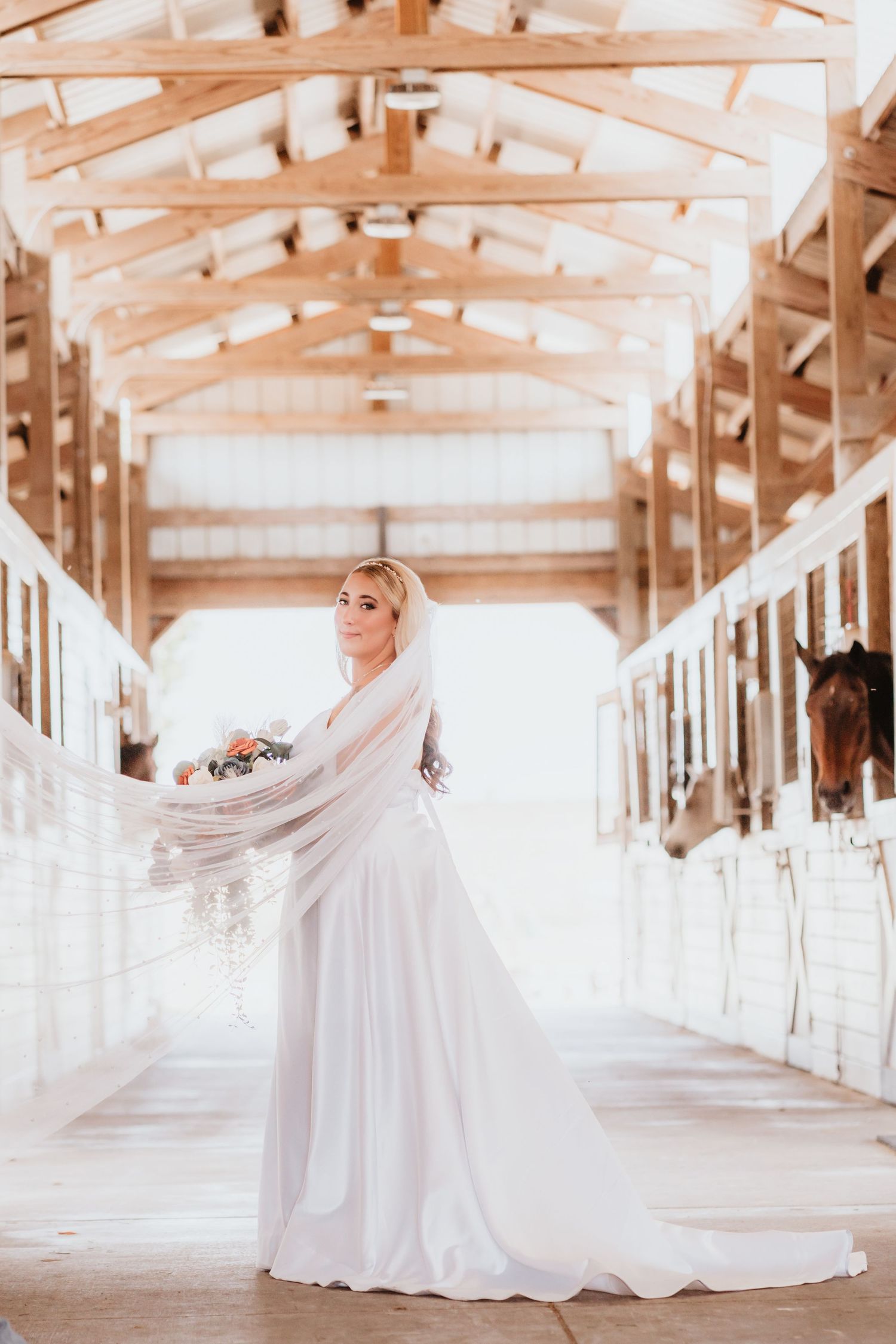 Bride in a white gown smiles while her veil flows in a bright horse stable with stalls on both sides.