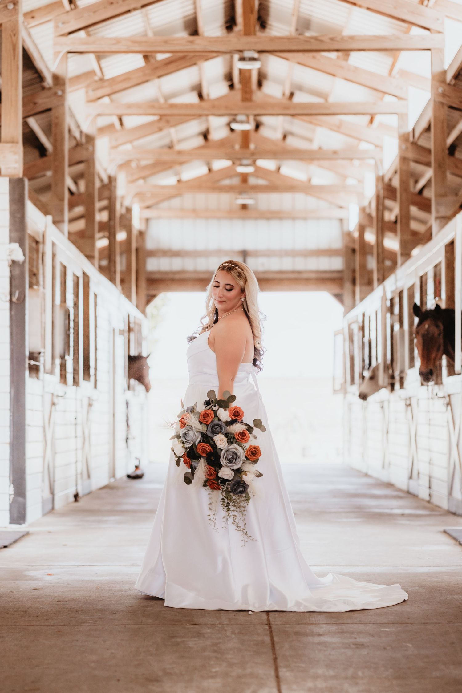 Elegant bride in a white gown holds a floral bouquet while standing in a sunlit horse stable aisle.