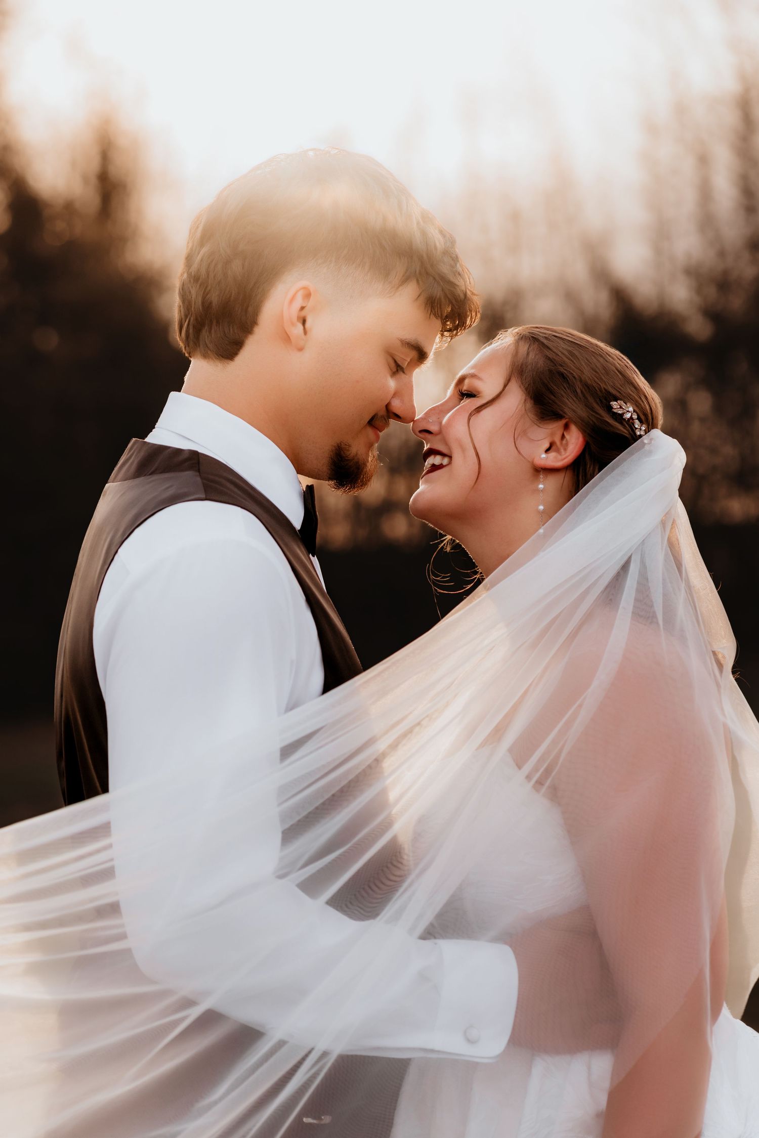 Bride and groom share romantic moment under flowing veil in warm golden sunset light.