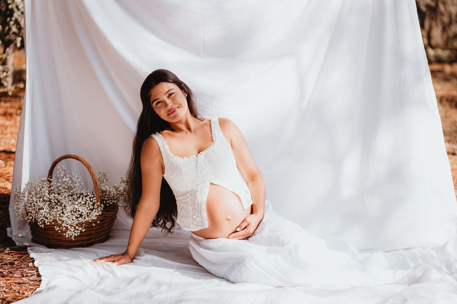 Serene pregnant woman in white outfit seated outdoors against flowing white fabric with flower basket.