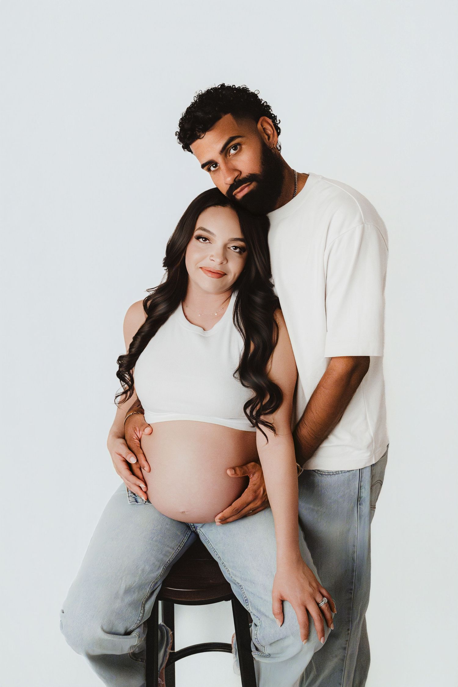 Expecting couple in white tops and jeans, man resting head on pregnant woman's shoulder on stool.
