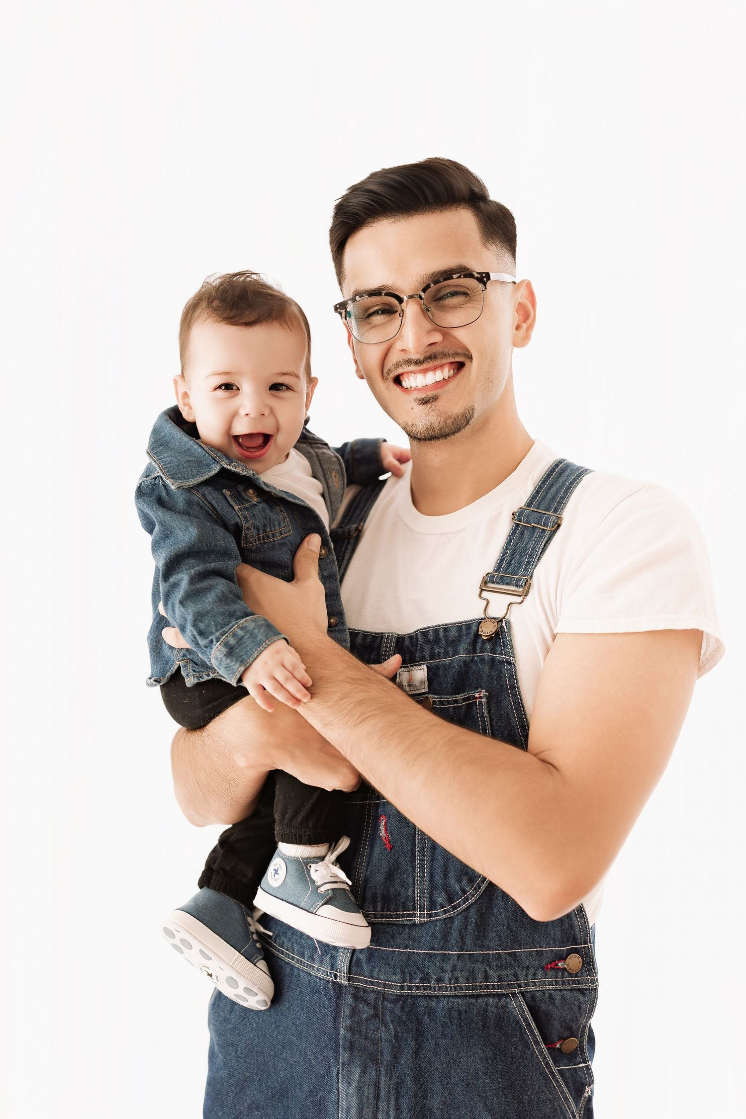 Smiling young father in denim overalls holding his laughing baby dressed in a denim jacket.