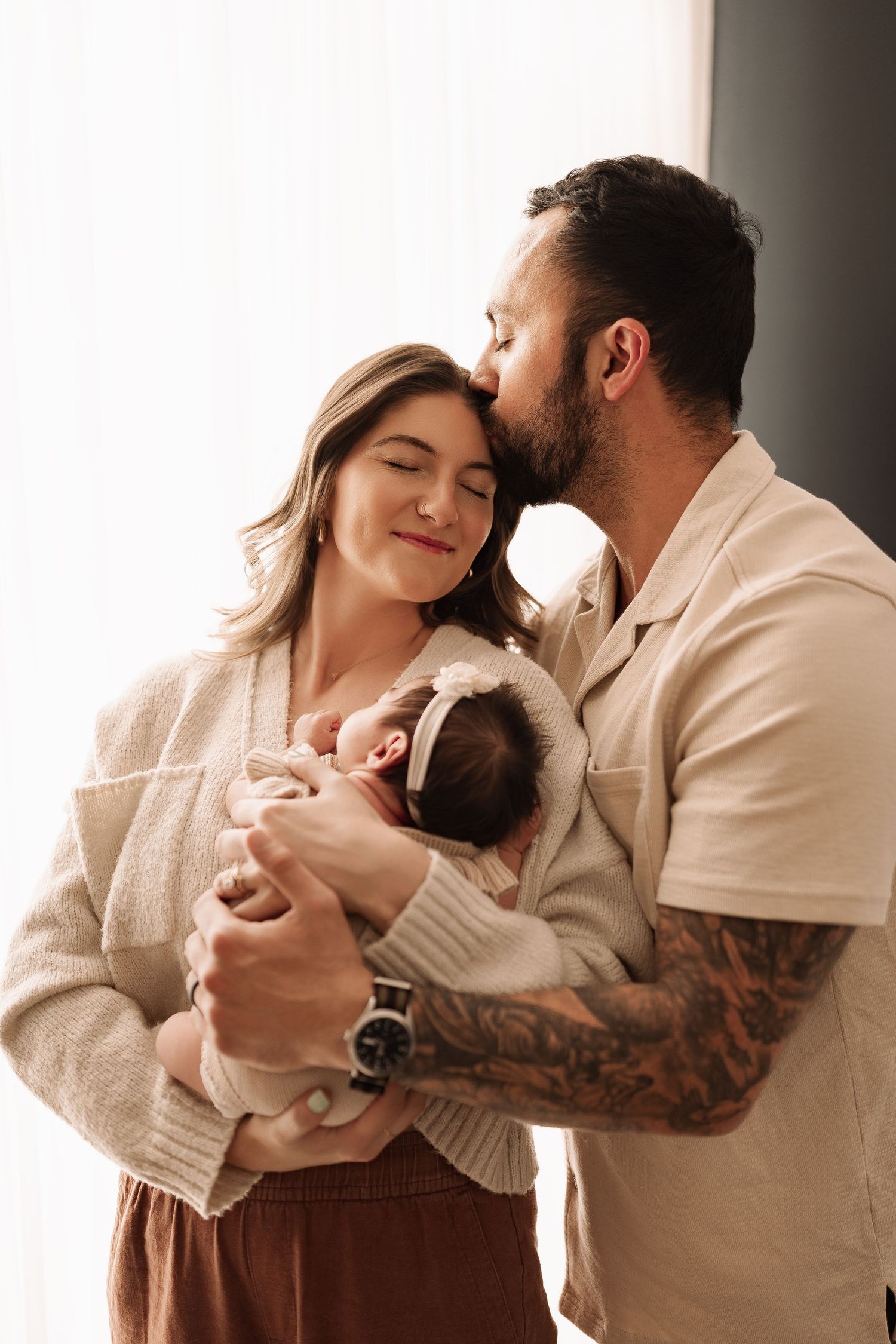 A bearded father kisses his smiling wife on the forehead as they both hold their newborn baby girl.