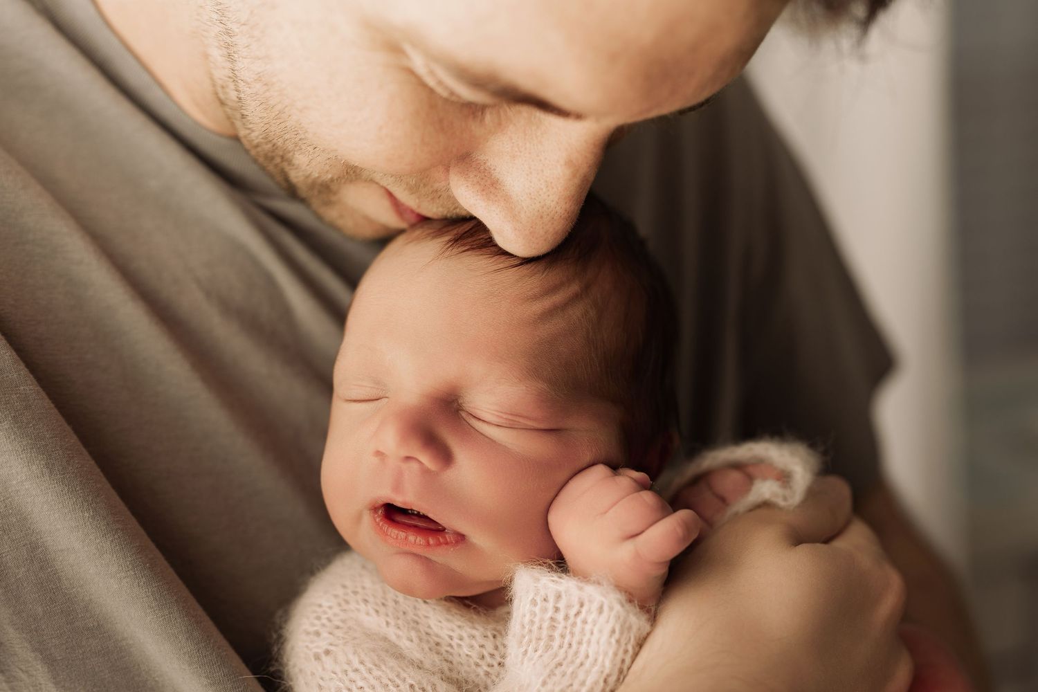 Father gently kissing his sleeping newborn baby wrapped in a soft knit blanket.