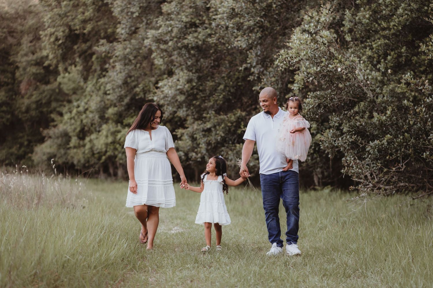 Happy family of four walking together through a green meadow surrounded by trees.
