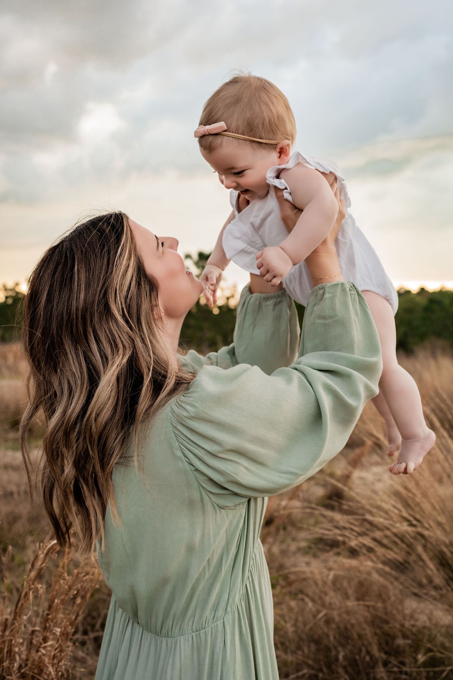 Mother in sage green dress lifting laughing baby girl overhead in golden grass field at sunset.