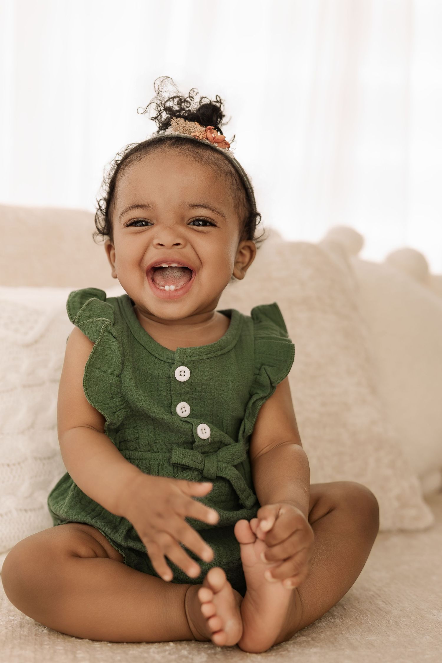 Smiling baby girl in green ruffle romper sitting cross-legged on white bed with floral hair accessory.