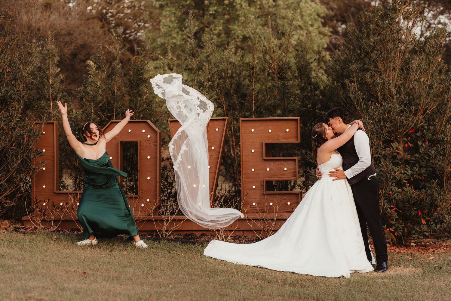 Bridesmaid joyfully tossing wedding veil as bride and groom kiss by lit LOVE marquee sign.