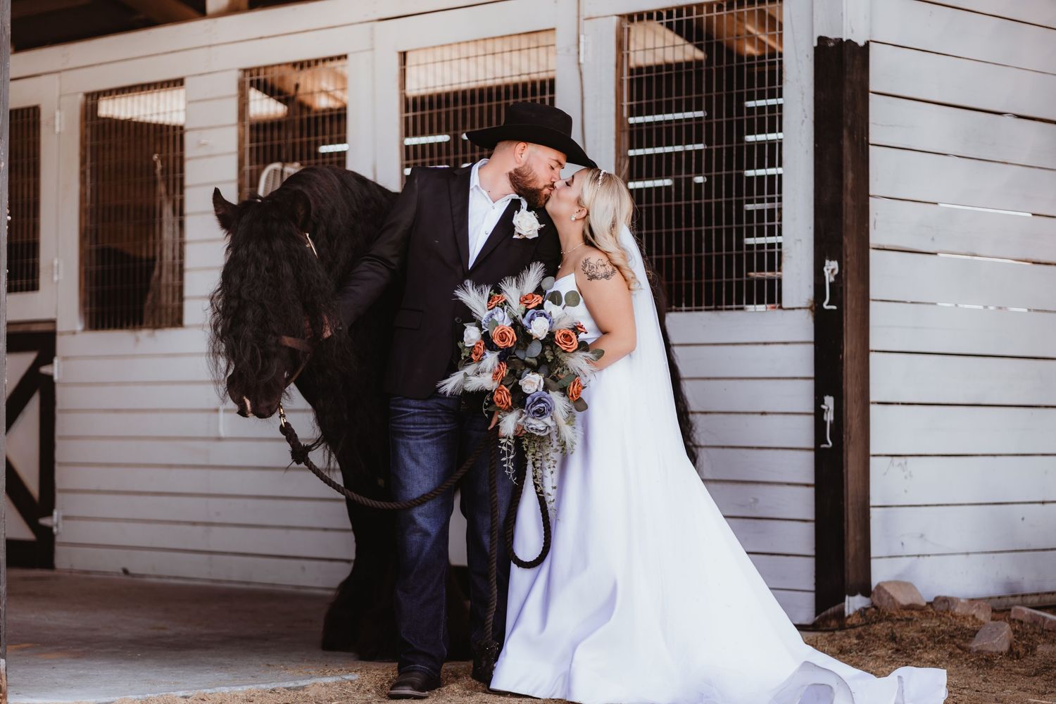 Bride and groom embrace with a black horse at a rustic barn wedding.