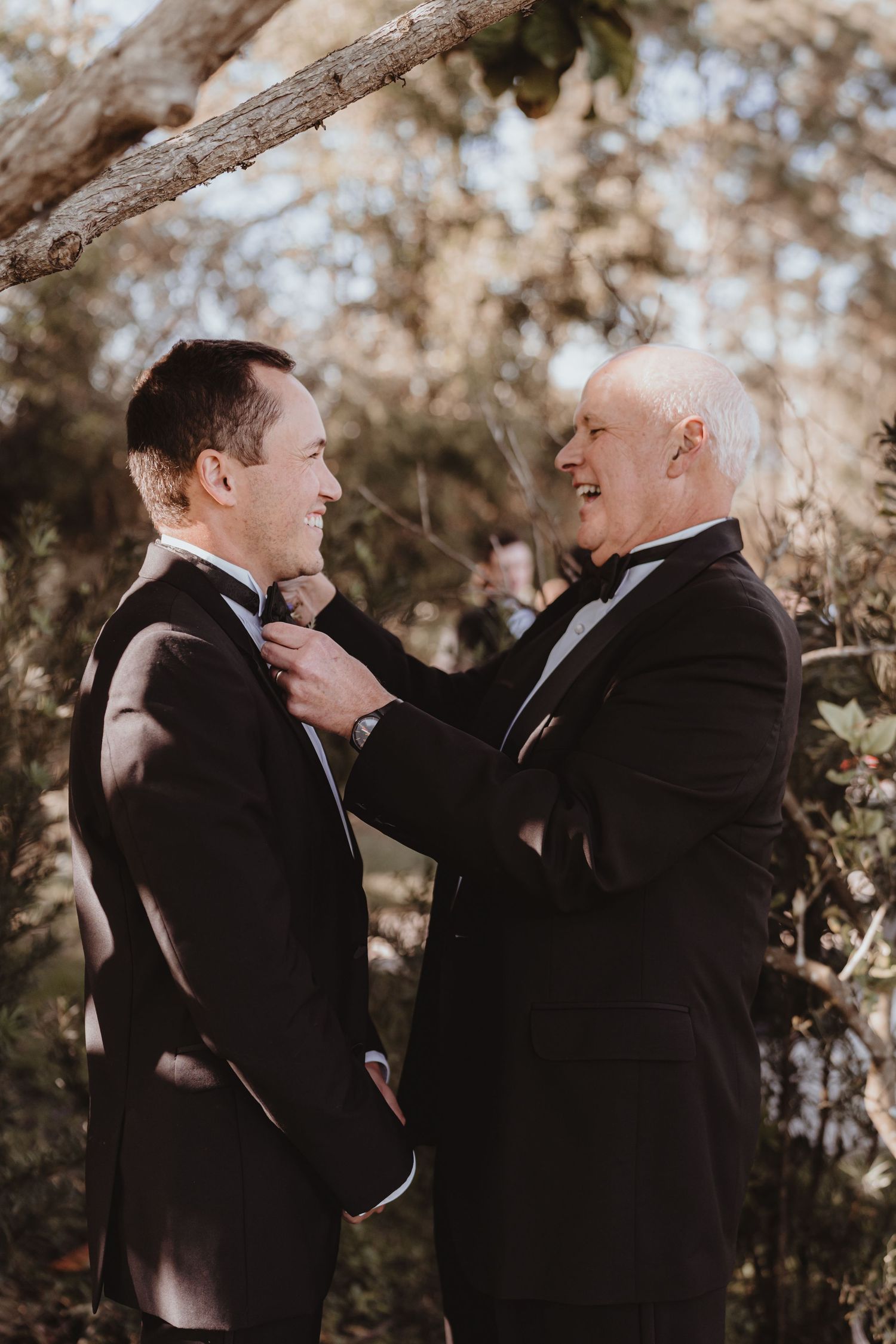 Older man in tuxedo adjusting groom's bow tie under a floral arch before the wedding ceremony.
