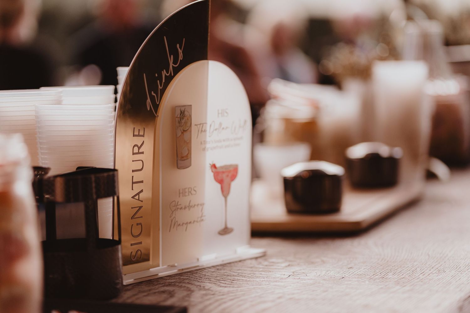 Wedding signature drinks menu sign displayed on table with candles and glassware at reception.