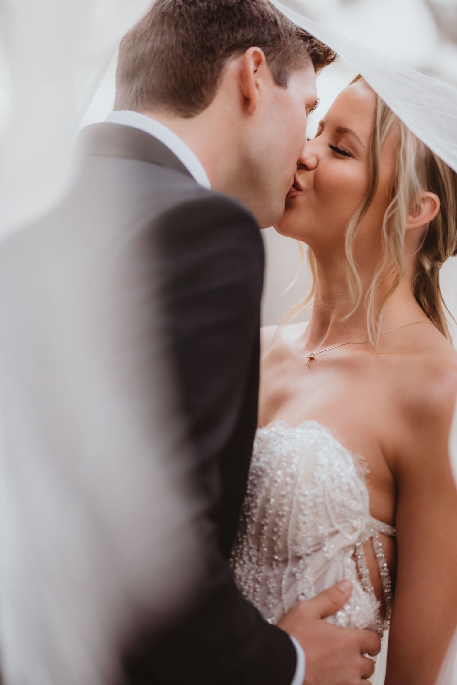 Bride and groom share a soft kiss beneath a flowing veil in romantic close-up.