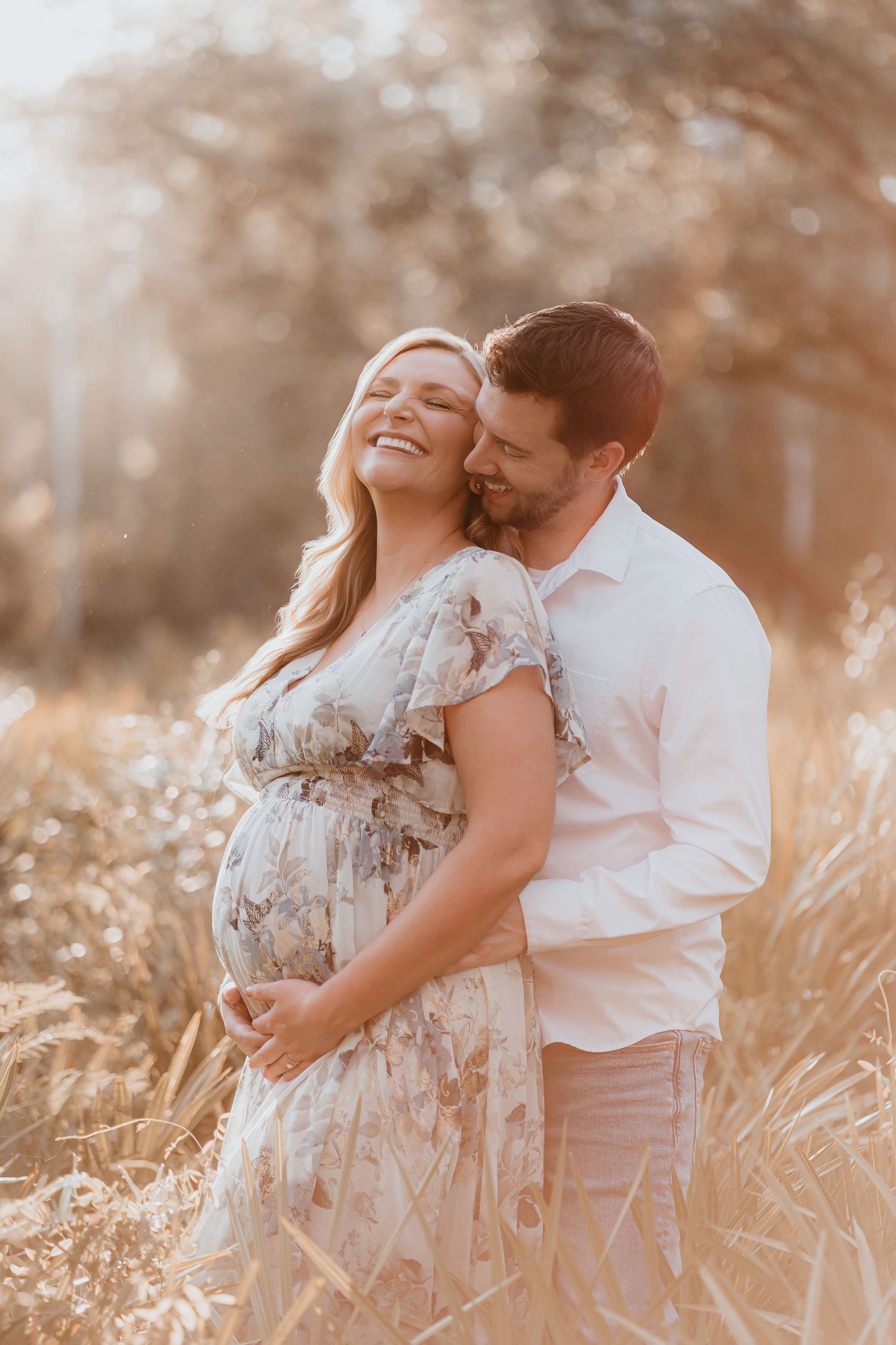 Pregnant woman in floral dress laughs joyfully as partner embraces her from behind in golden field.