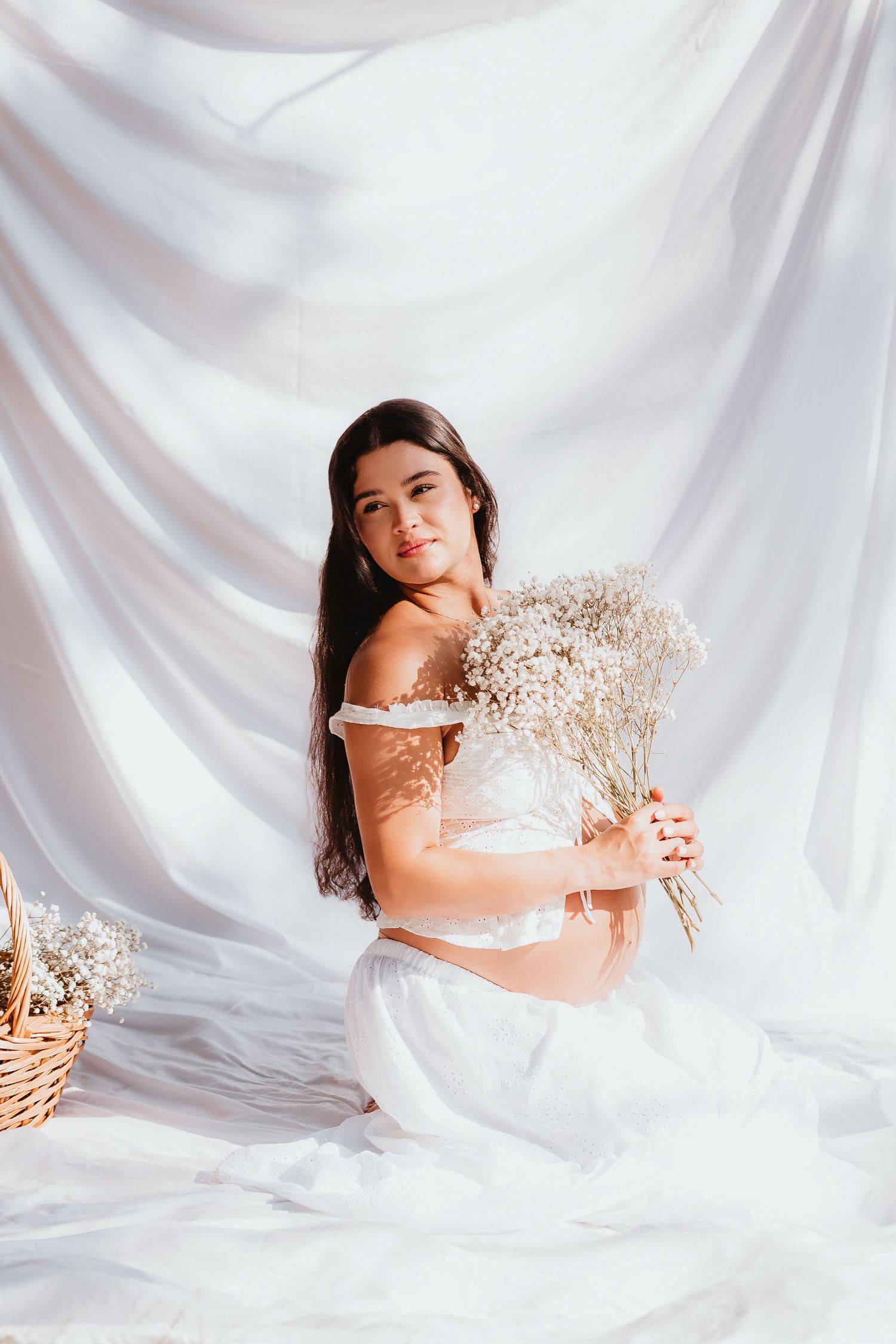 Pregnant woman in white off-shoulder dress holding baby's breath bouquet against white drape.