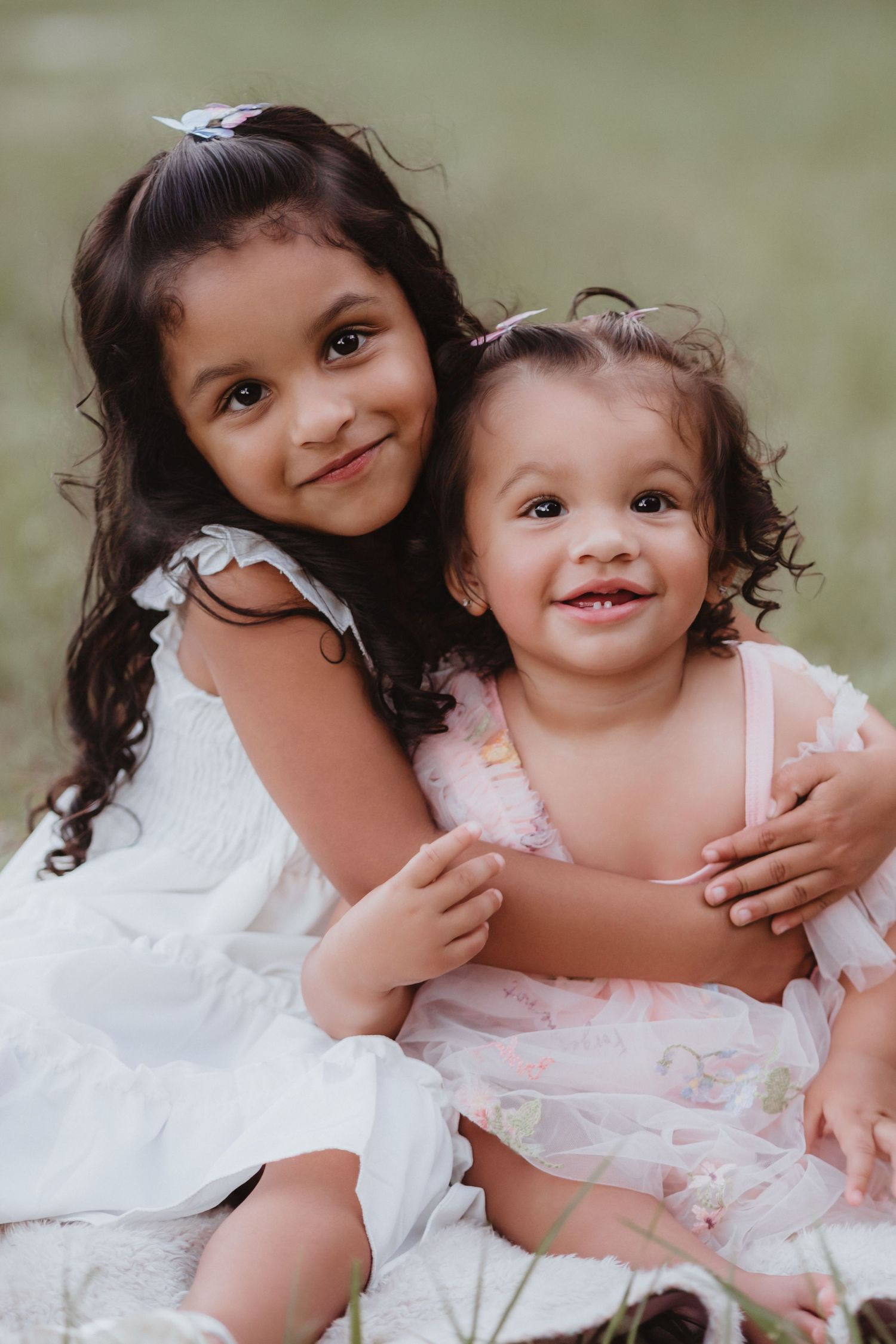 Two little sisters in white and pink dresses hugging and smiling at the camera outdoors.