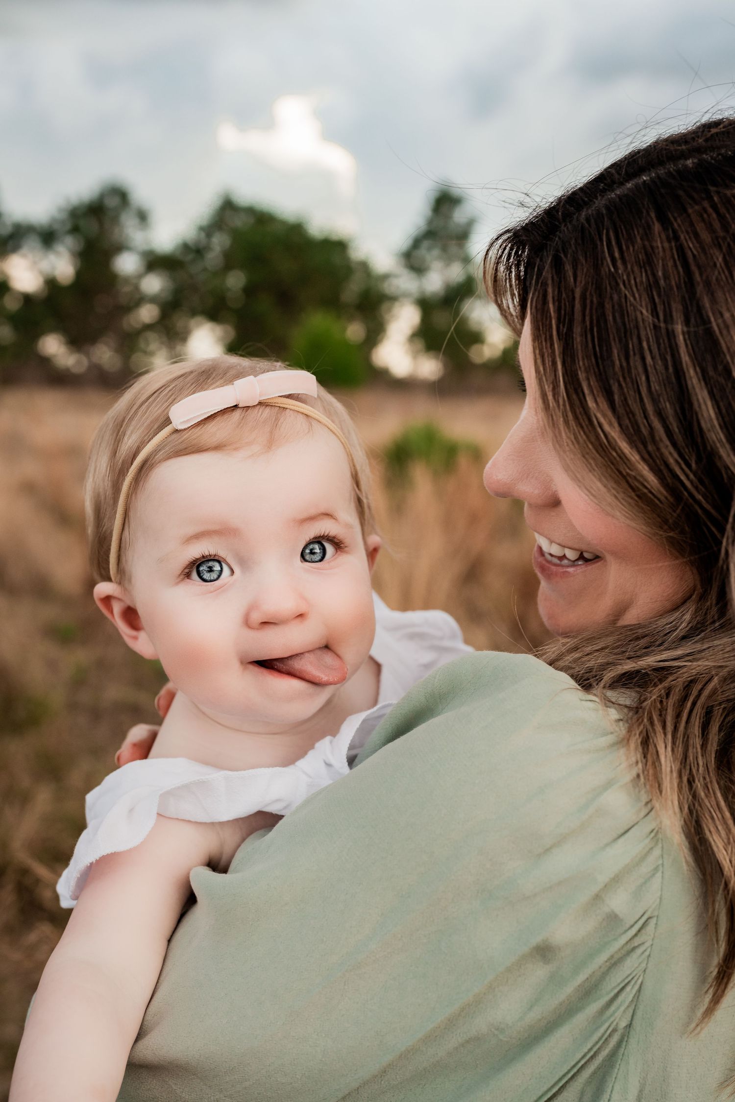 Adorable blue-eyed baby girl sticking out tongue while being held by smiling mother outdoors.
