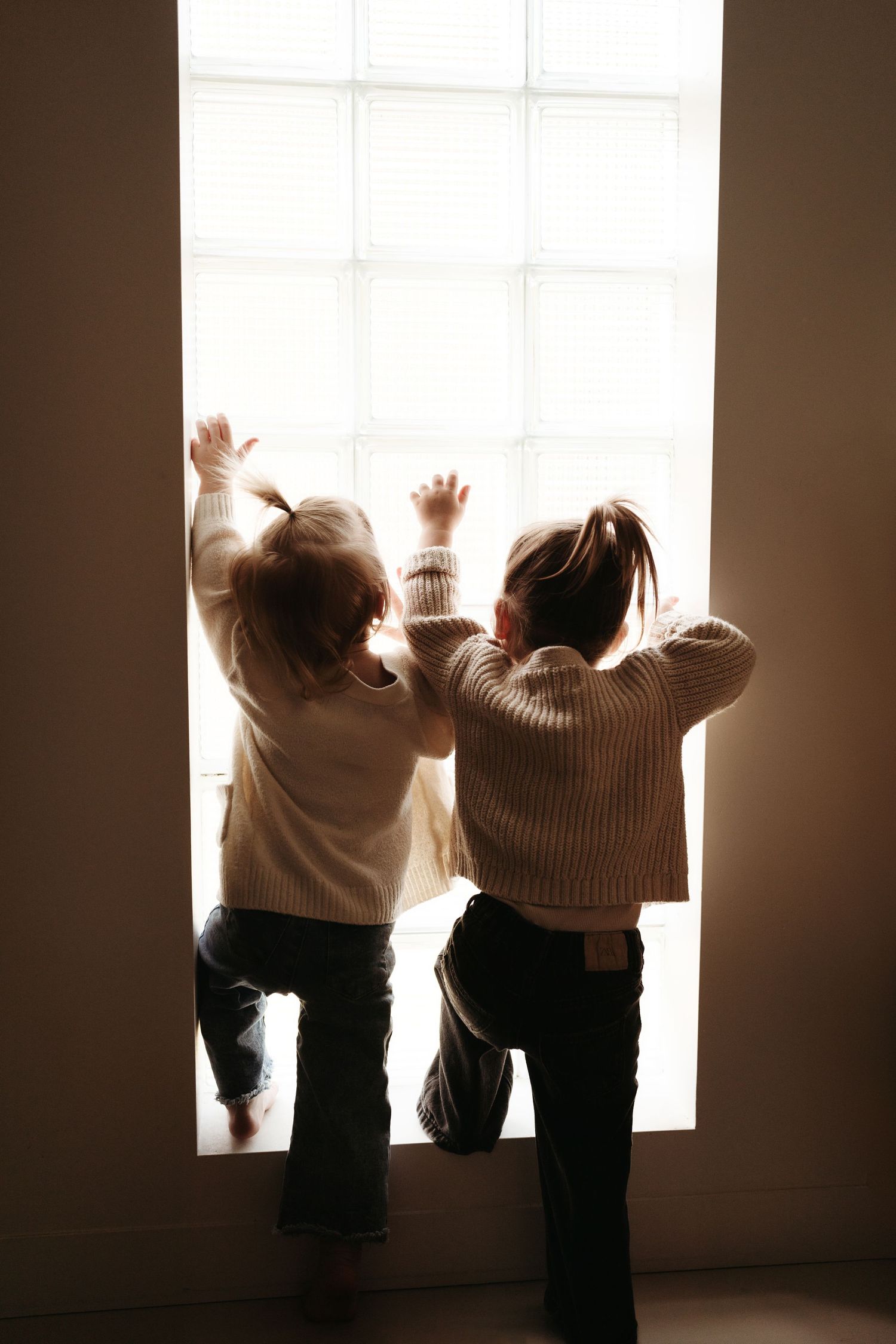 Two young siblings in matching beige sweaters reach up together toward a bright window.
