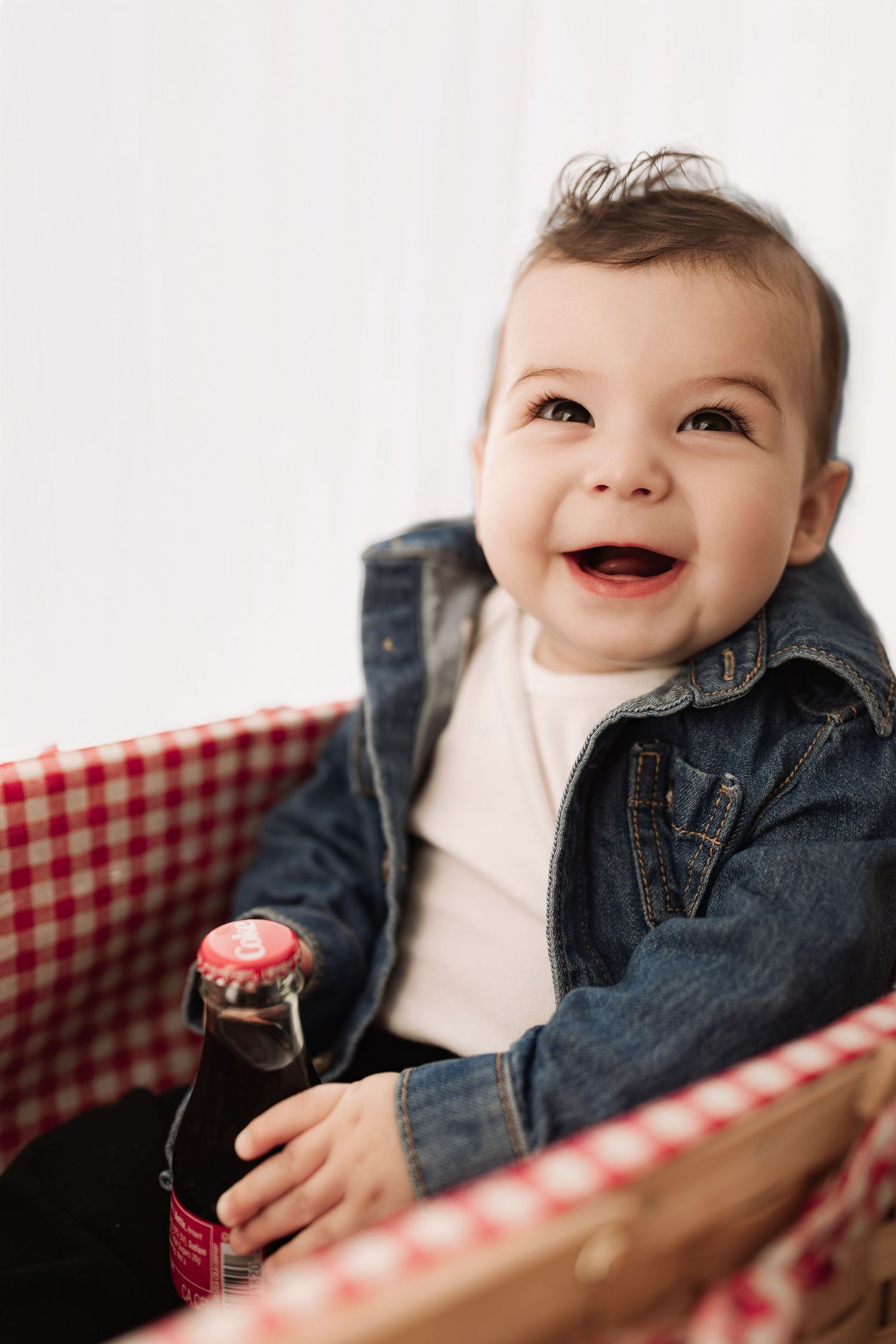 Adorable baby in denim jacket laughing while holding a small soda bottle in a picnic basket.