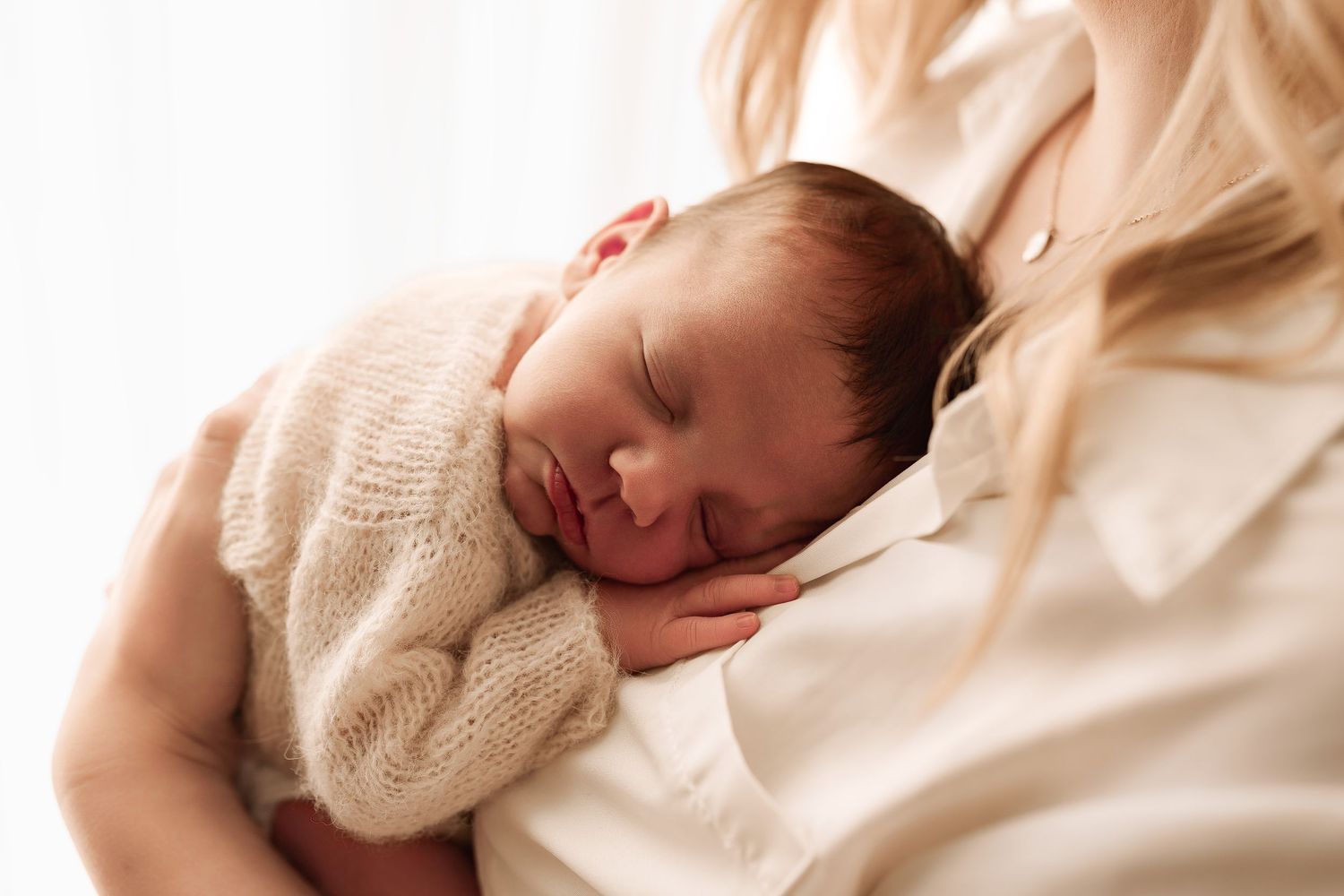 Mother cradling her sleeping newborn baby dressed in a soft cream knit outfit.