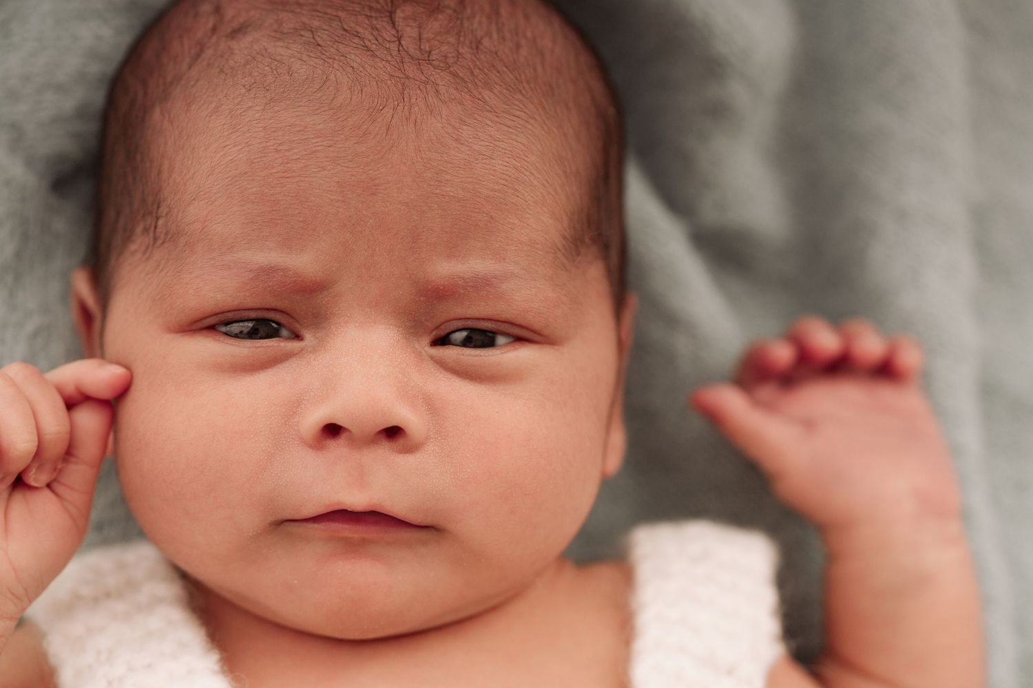 Close-up portrait of a newborn baby with dark hair lying on a gray blanket, looking alert with tiny hands raised.