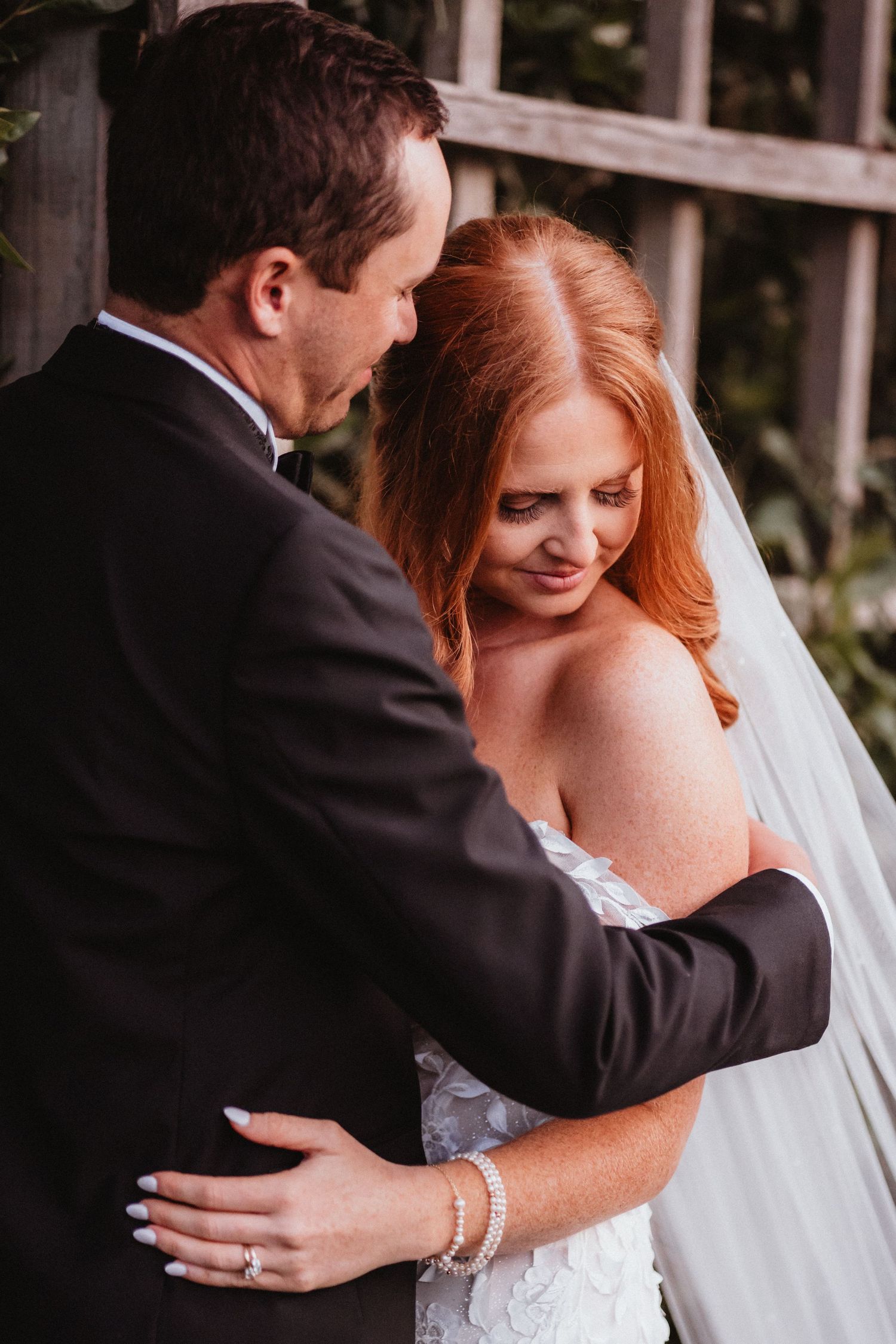 Groom embraces bride in floral wedding gown against a wooden trellis.