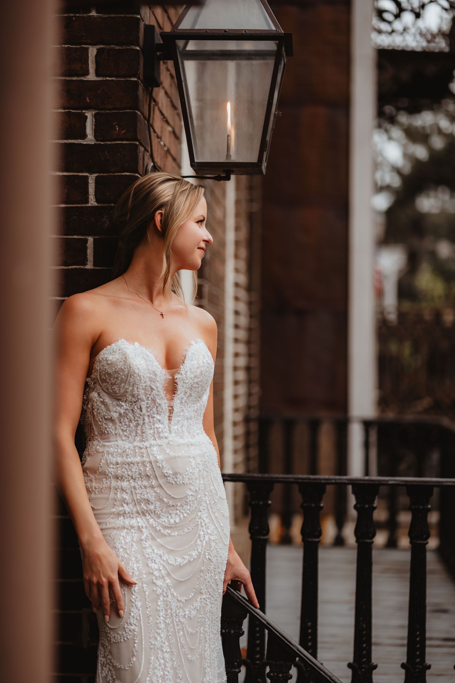 Elegant bride in strapless lace gown poses on a brick porch beside a vintage gas lantern.