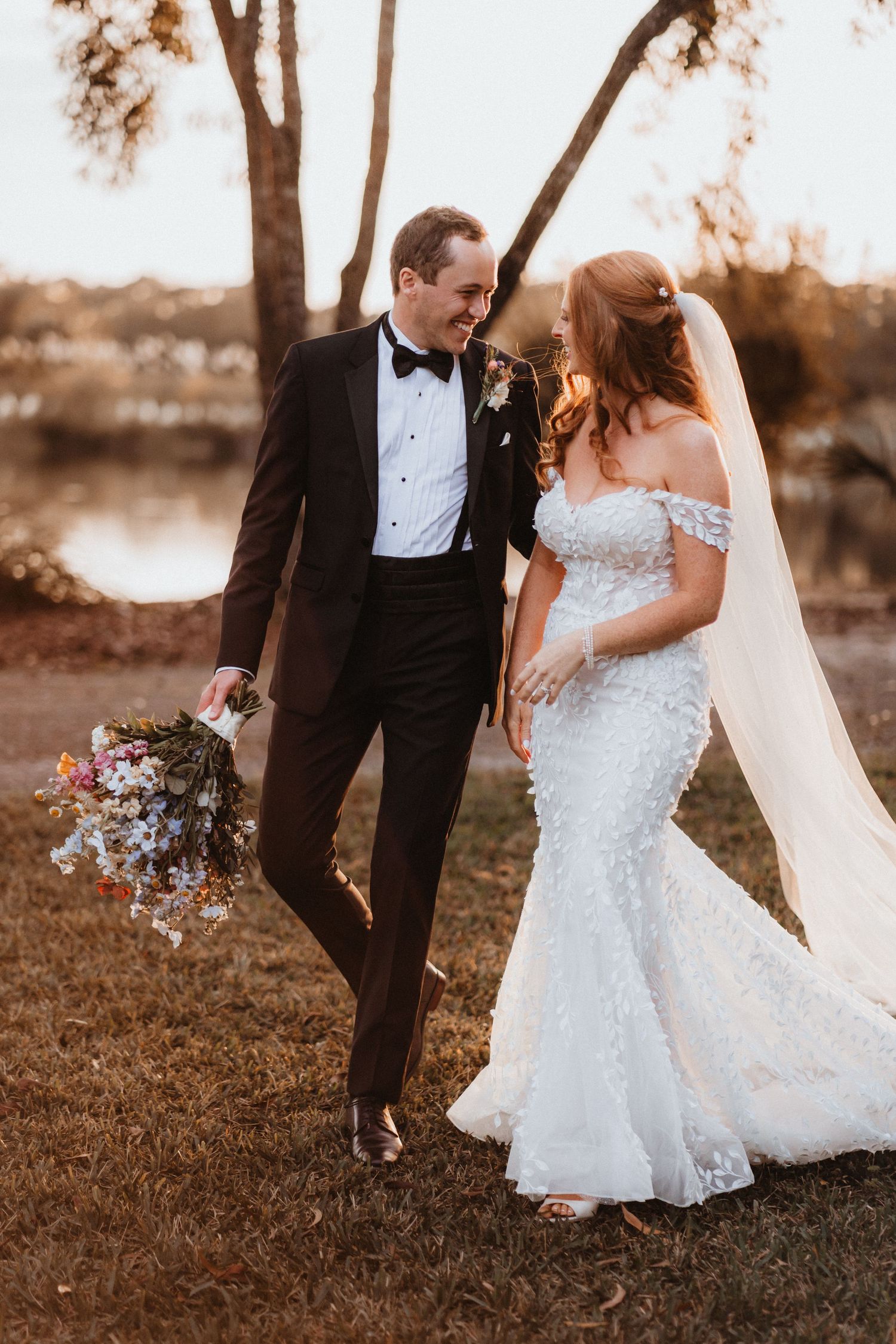 Bride and groom walk together outdoors at golden hour, she in lace mermaid gown, he in black tuxedo holding bouquet.