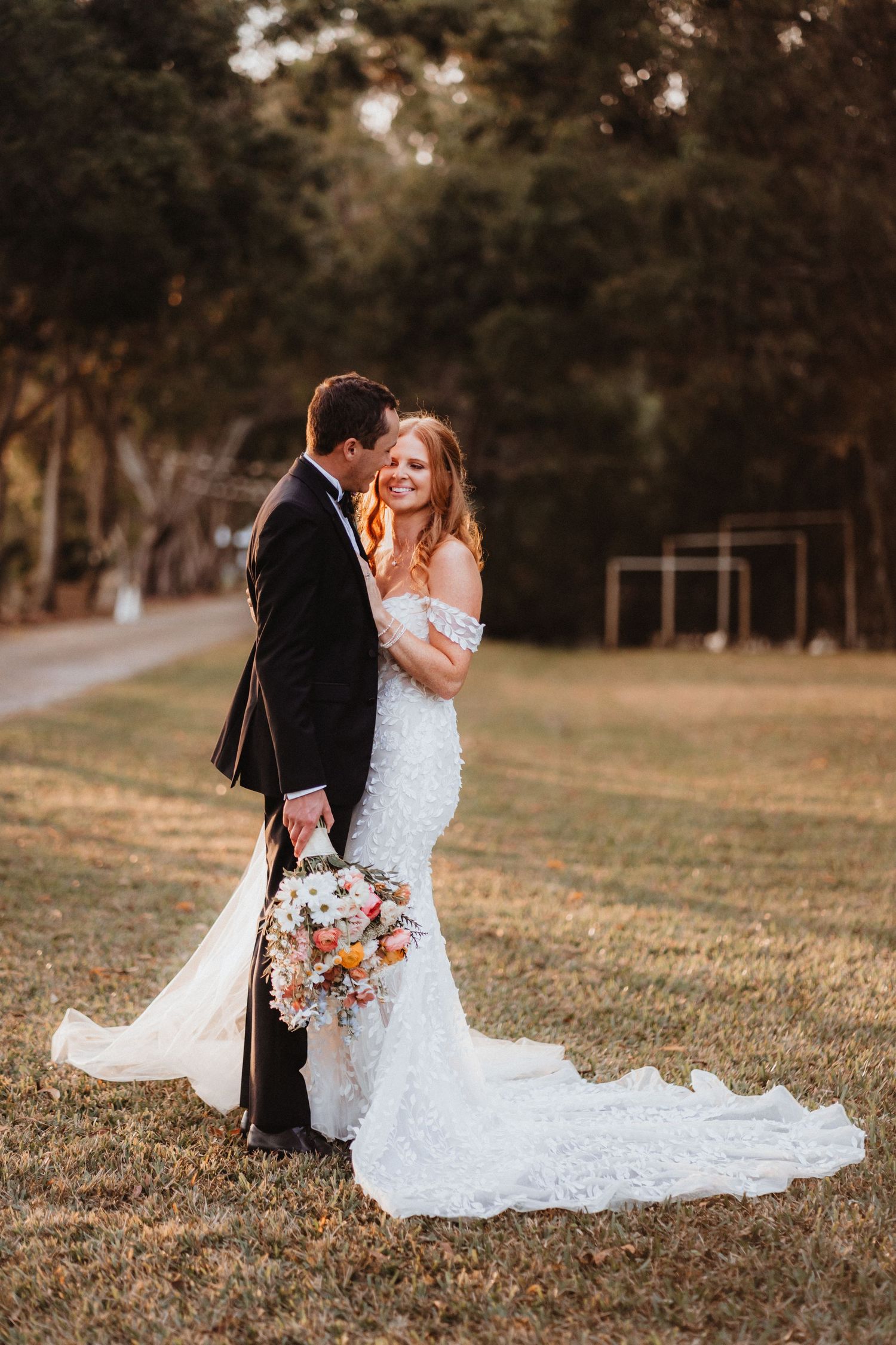 Groom kisses bride on cheek as she holds colorful bouquet in off-shoulder lace gown during sunset portrait.