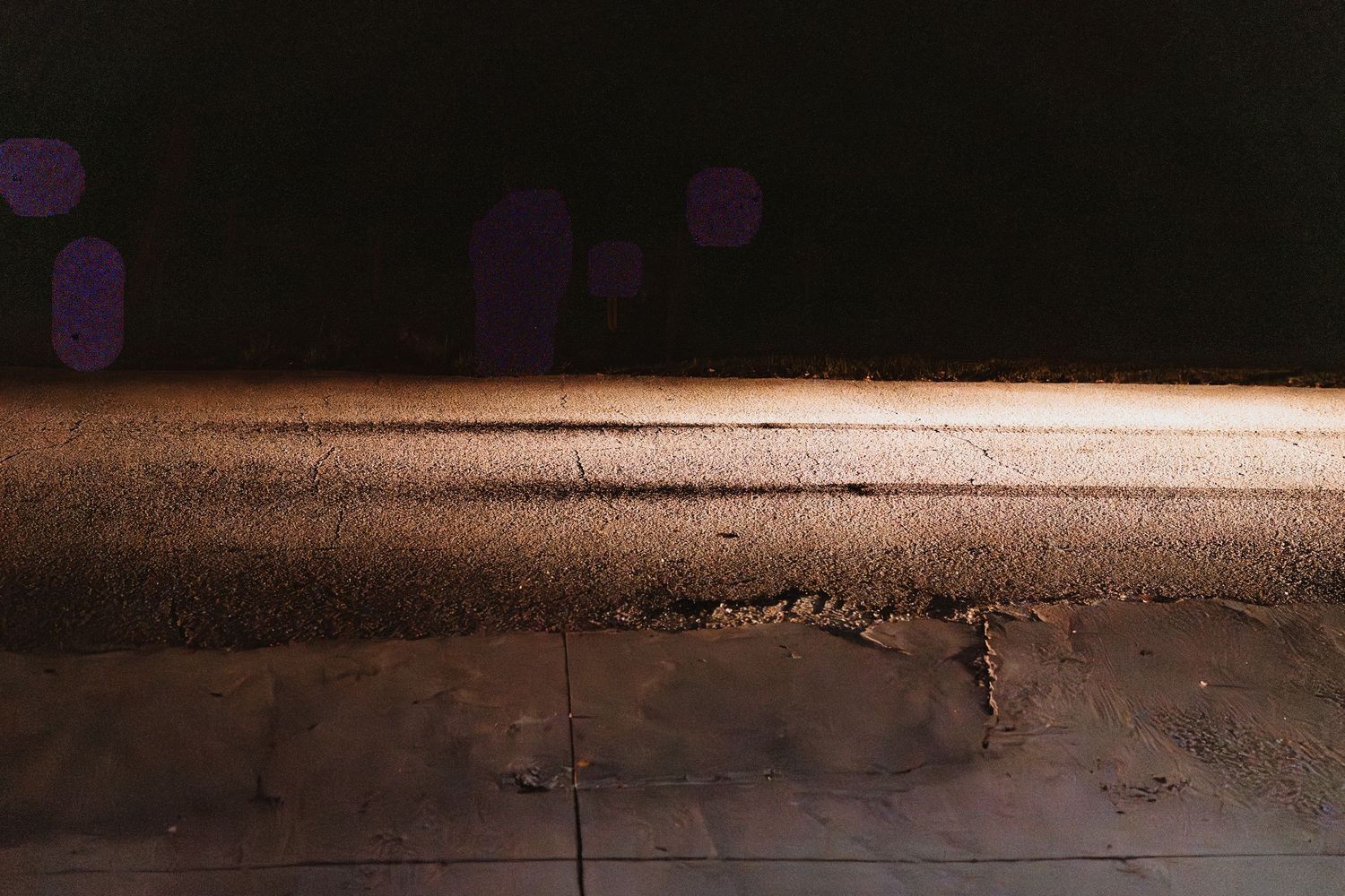 Close-up of weathered concrete curb edge with purple light bokeh reflections on dark pavement at night.