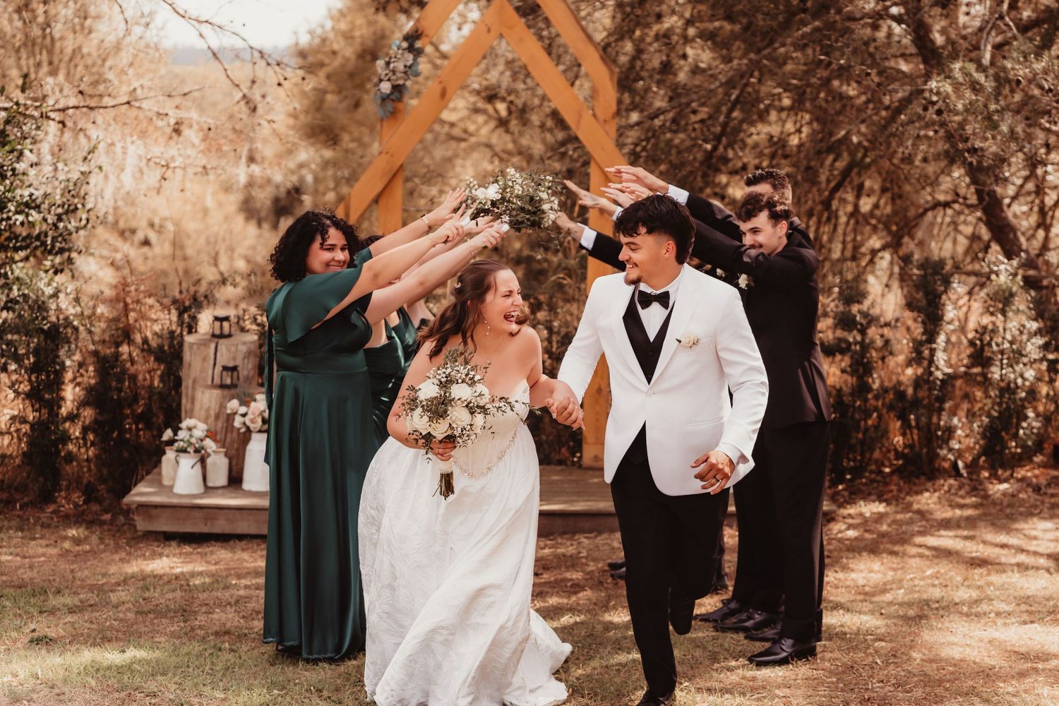 Newlyweds holding hands walking joyfully as wedding party celebrates with raised hands outdoors.