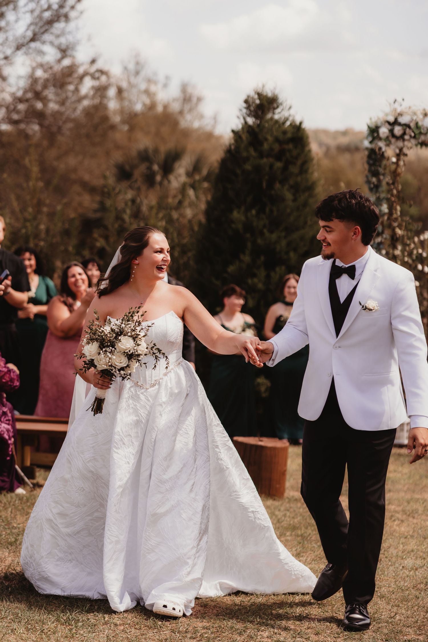 Happy newlyweds walking down aisle, bride in white gown holding bouquet, groom in white tuxedo jacket.