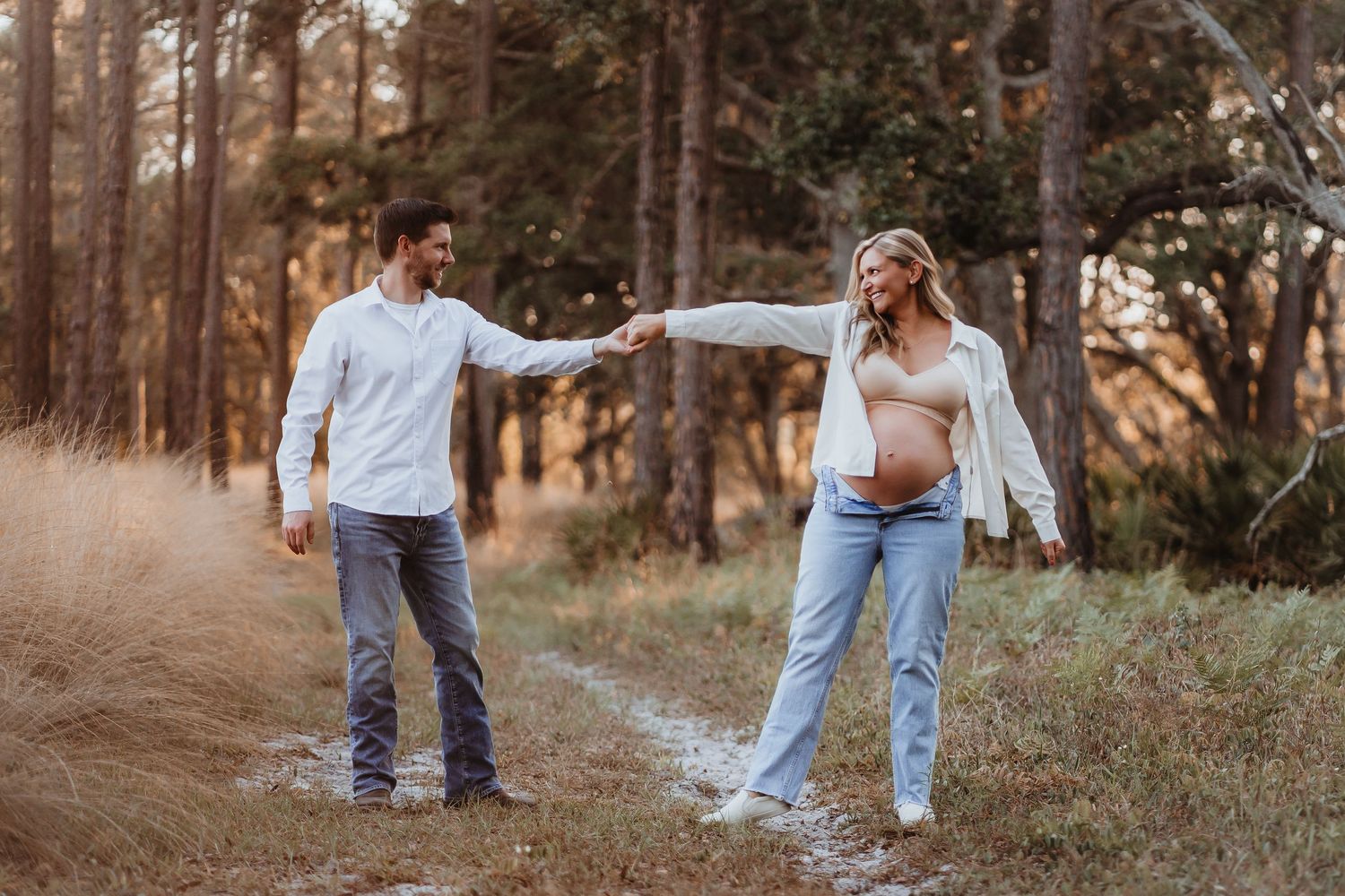 Pregnant couple holding hands and walking on a sunlit forest path.
