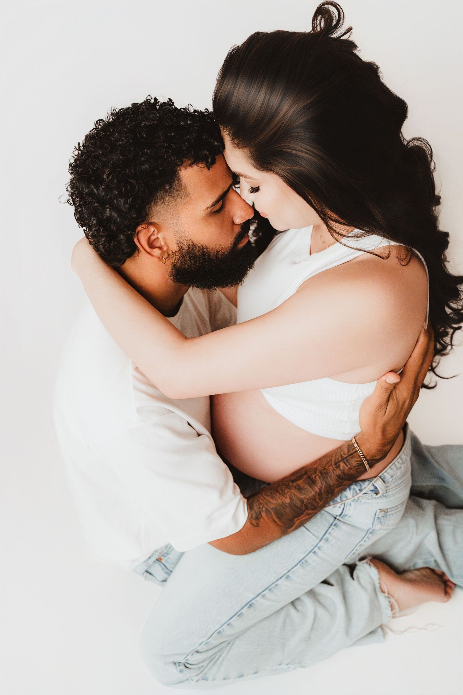 Man embracing pregnant partner from behind, both leaning in for a kiss in white studio.