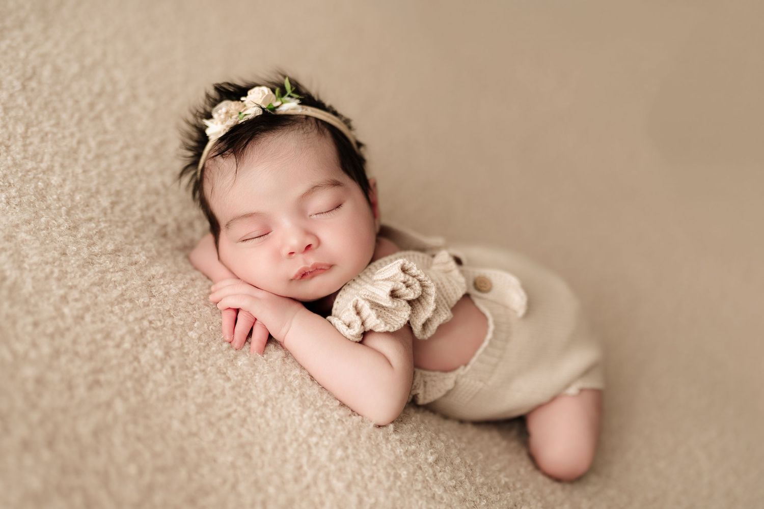 Sleeping newborn baby girl in beige romper and floral headband posed on soft tan blanket.