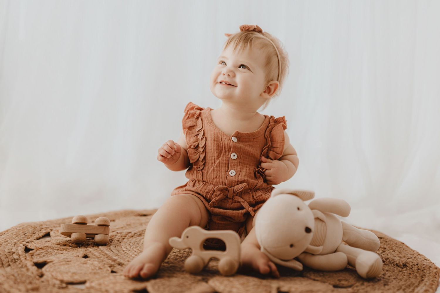Smiling baby girl in rust romper sitting on woven rug with stuffed bunny toy.