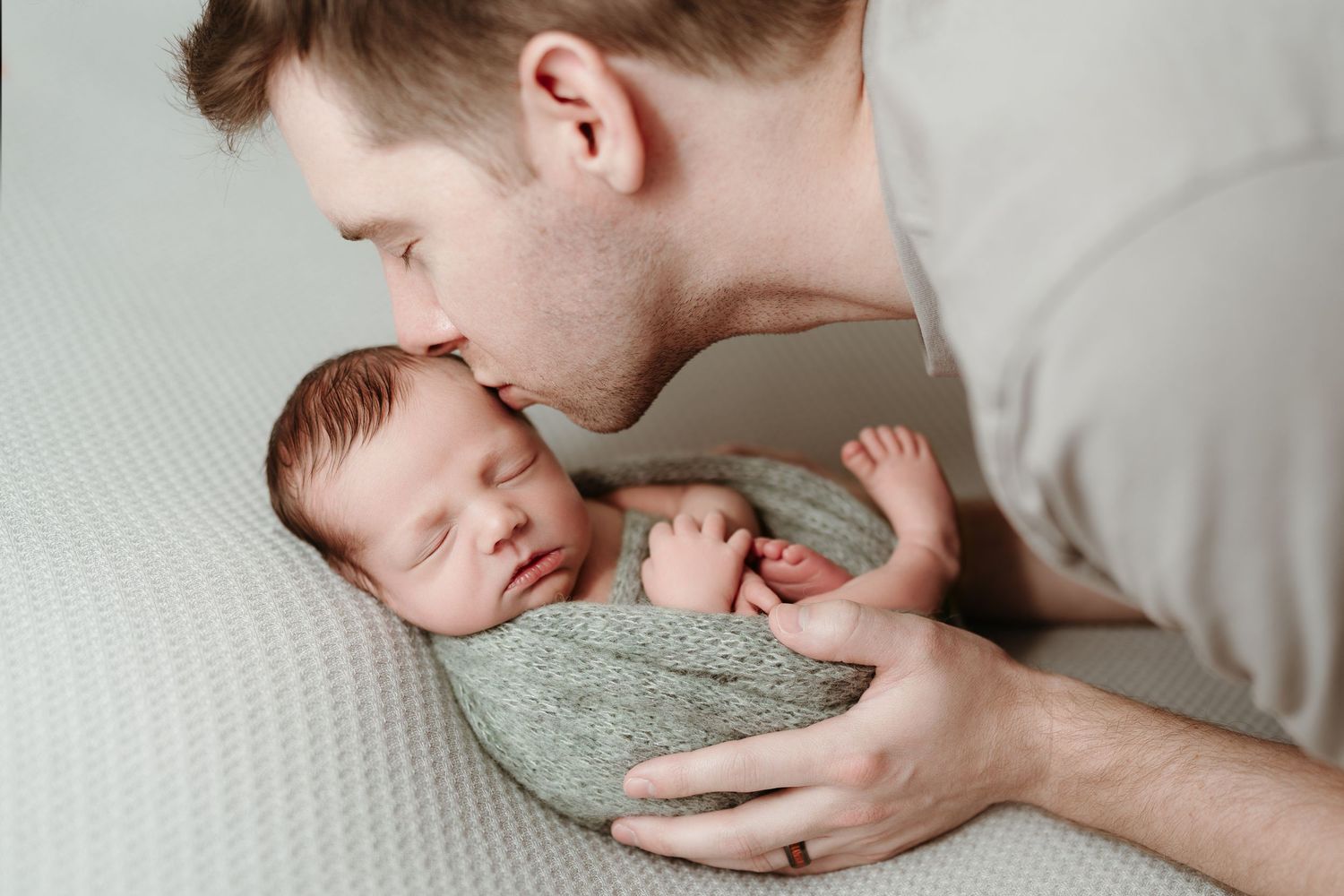 Father tenderly kisses sleeping newborn baby wrapped in green knit blanket.