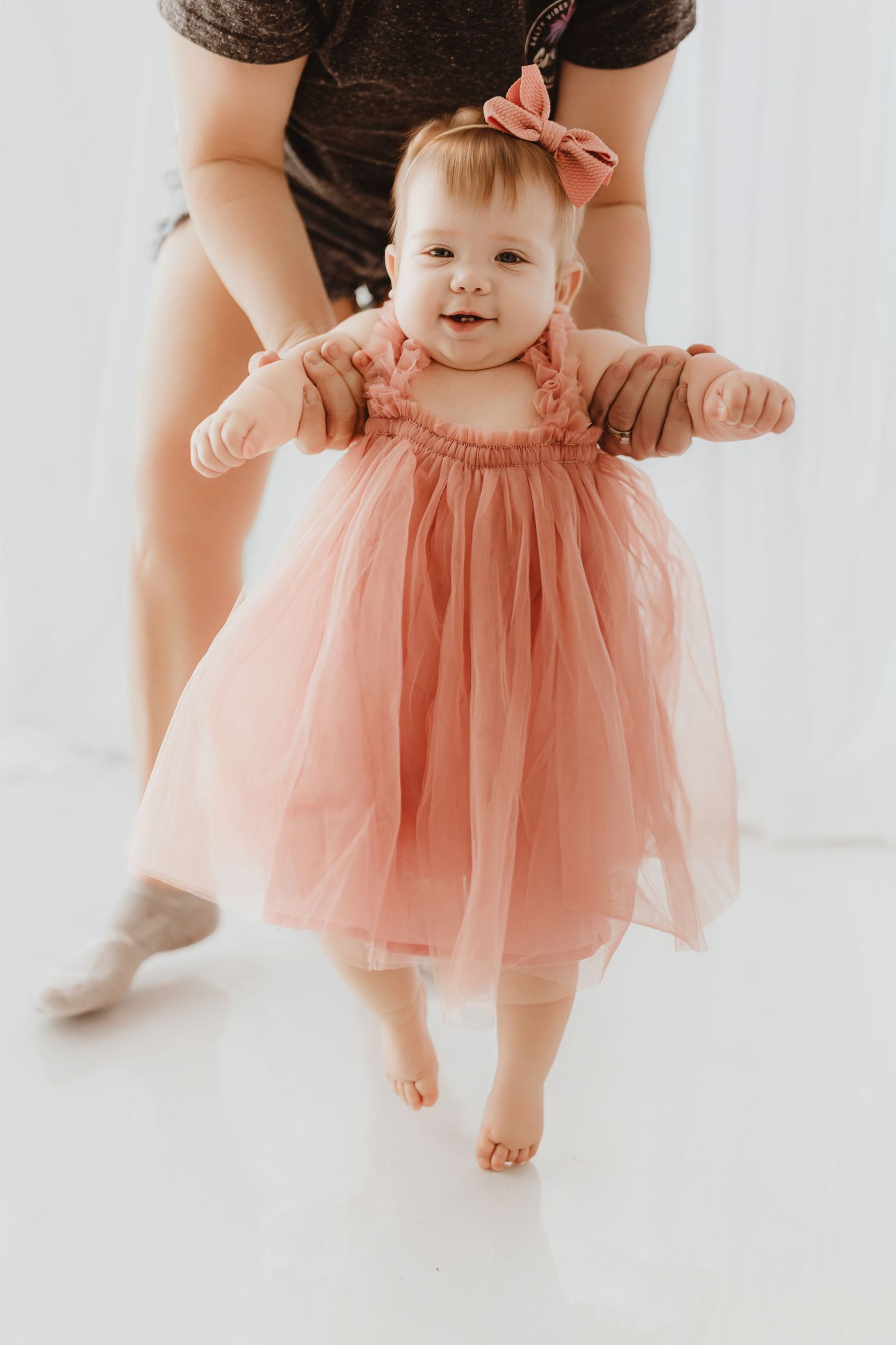 Adult helping baby girl in pink tulle dress and bow headband practice her first steps in studio.
