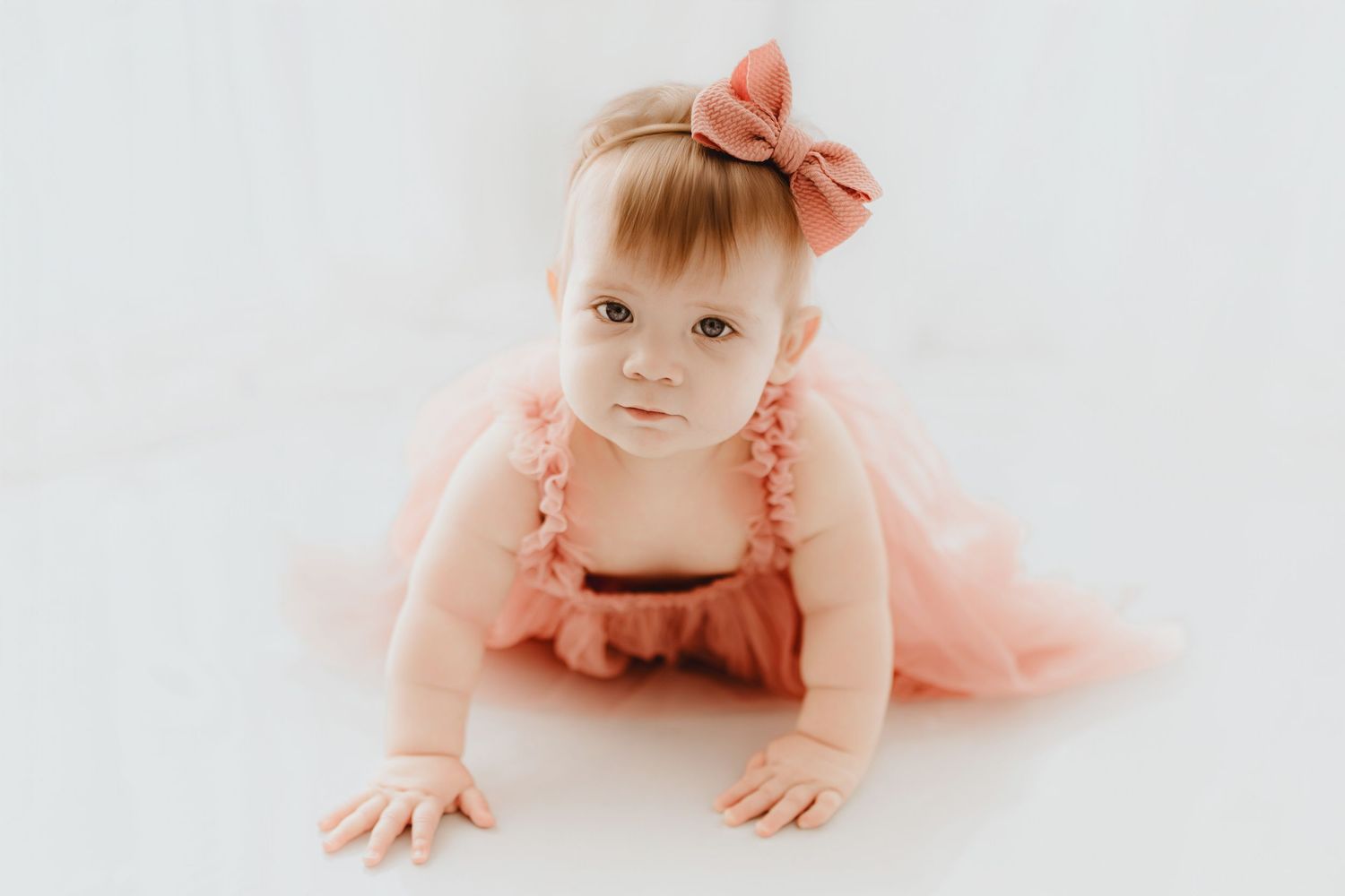 Adorable baby girl in pink ruffle romper and bow headband crawling on white background.