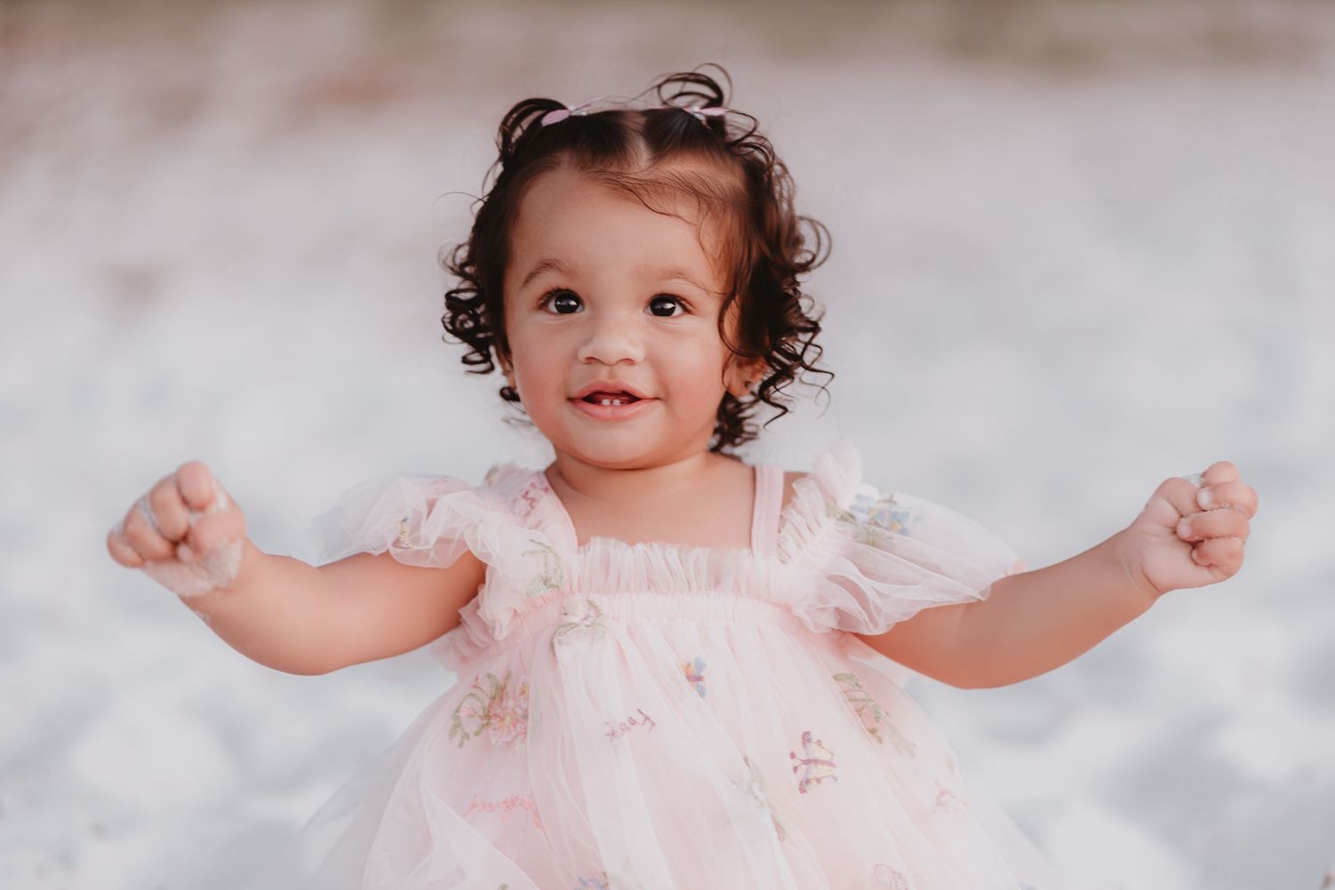 Happy toddler girl in pink floral dress with curly hair smiling with arms outstretched on beach.