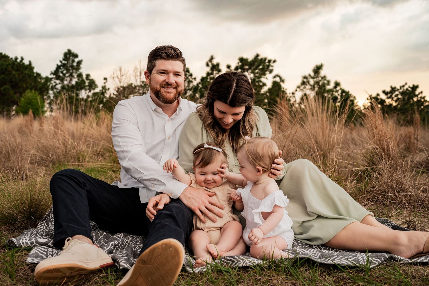 Family of four smiling together on a blanket in a golden field with two baby girls at sunset.