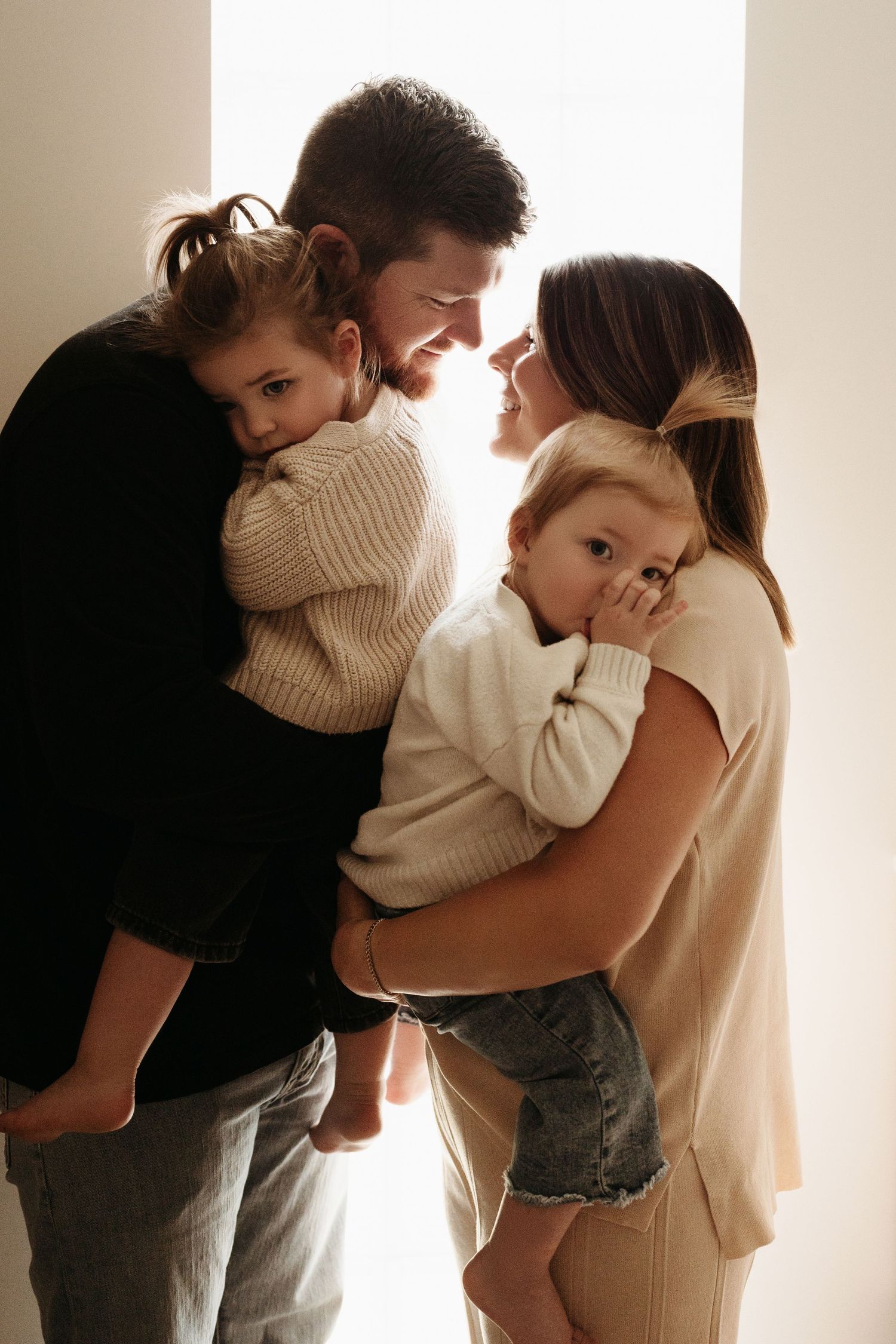 A happy family of four embraces near a bright window, parents each holding a young child.