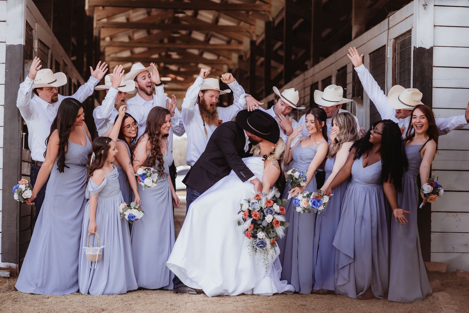 Wedding party celebrates with raised arms as bride and groom share a kiss, bridesmaids in lavender dresses.